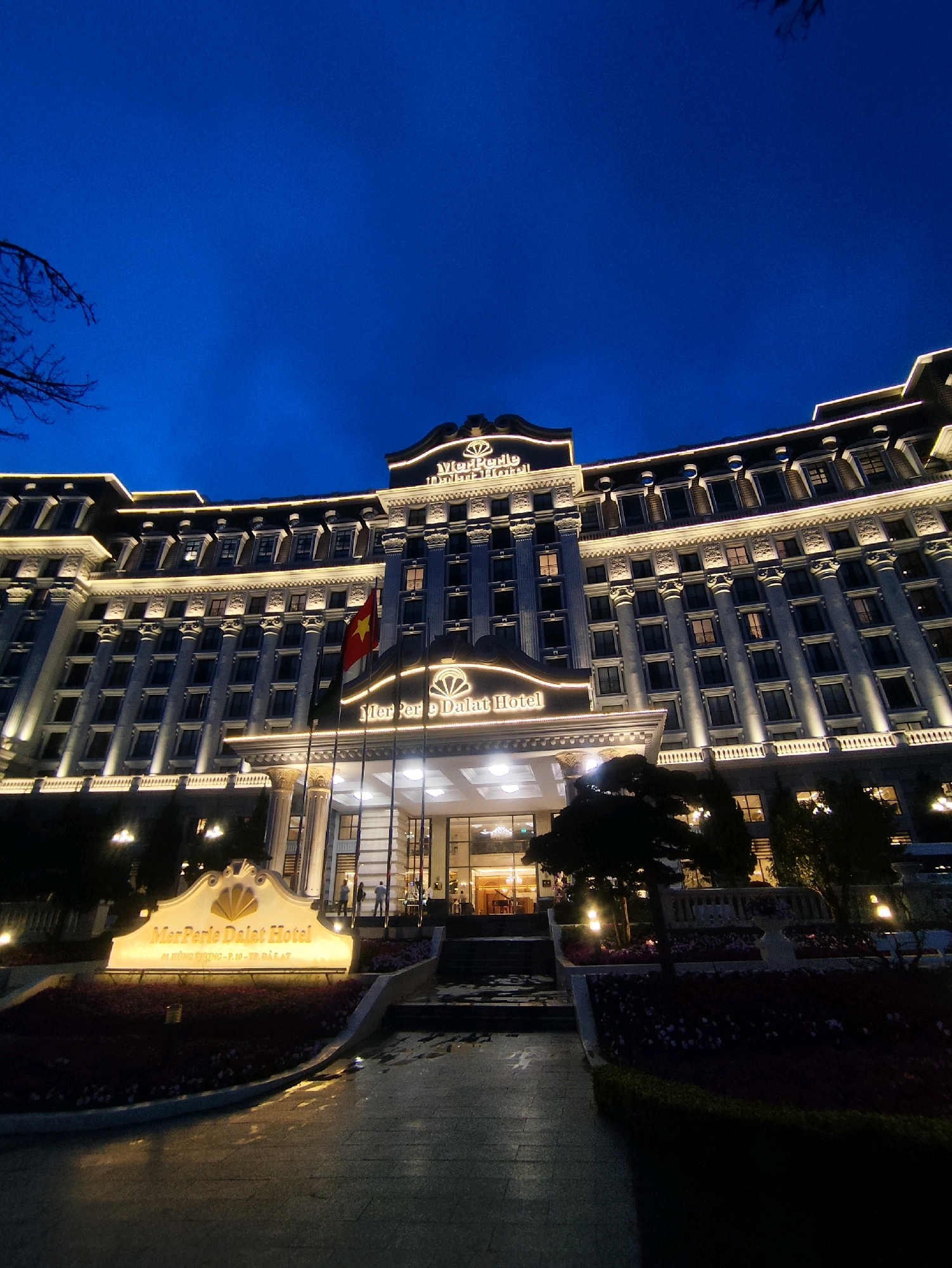 The image shows the MerPerle Dalat Hotel in Da Lat, Vietnam, at night. The hotel is a large, ornate building with many windows and decorative columns, illuminated by exterior lighting. A prominent sign in front of the hotel reads "MerPerle Dalat Hotel" in illuminated letters. A Vietnamese flag is visible flying in front of the building. The scene is set against a dark blue night sky, with some trees and landscaping in the foreground. The ground in front of the hotel appears to be wet, suggesting recent rain or mist. There are a few people visible near the entrance of the hotel. The overall impression is one of a grand and well-lit establishment.