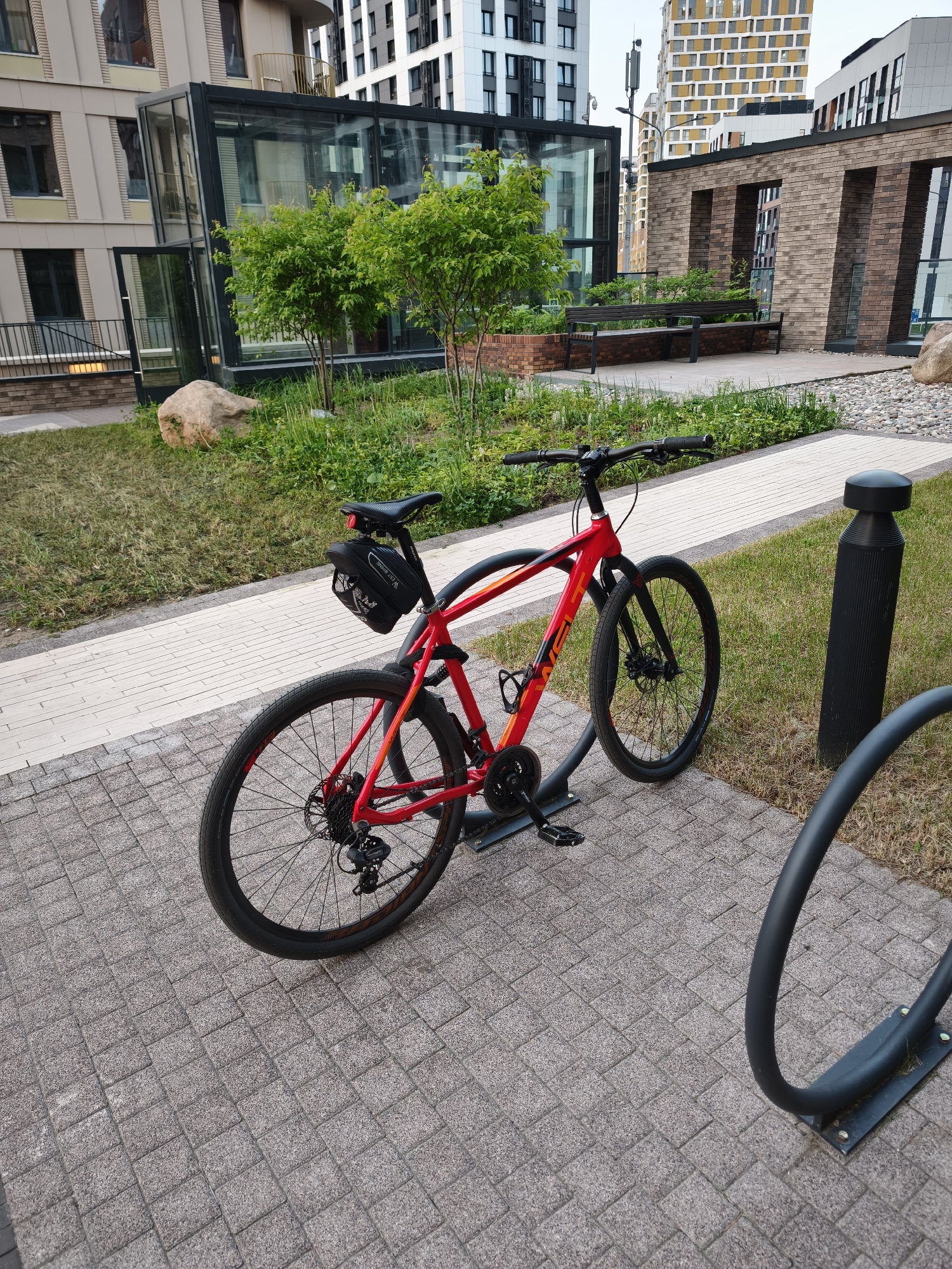 A red and black hybrid bicycle is secured to a gray metal bike rack on a paved area constructed with small, textured gray pavers. The bicycle has a black bag attached under the seat. The bike rack is next to a patch of grass. Behind the bike, there is a low-level border constructed of light-colored brick pavers. Behind the brick border there is a manicured lawn and various shrubs. The architecture behind the bicycle includes a single-story glass-enclosed room and several high-rise apartment buildings. The photograph was taken in Moscow, Russia.