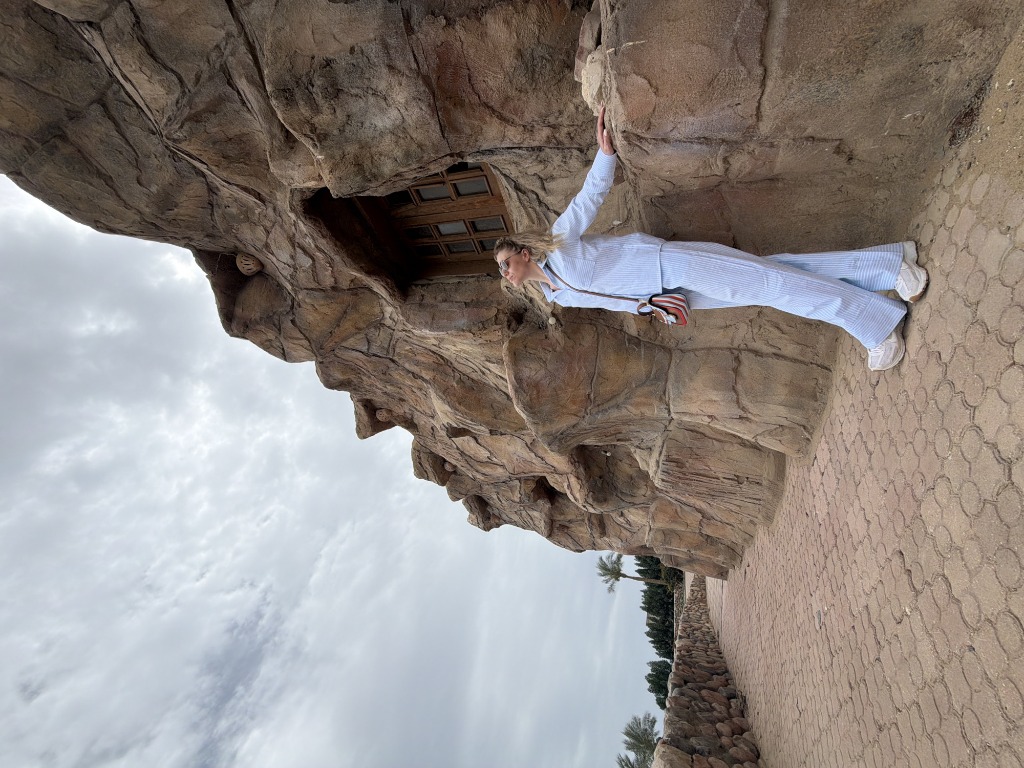 A woman in a light blue striped suit stands on a patterned stone walkway adjacent to a rock formation. She is positioned in profile facing left, with her right hand touching the rock face. The rock formation features a small window or opening with a grid-like design. Behind the formation, palm trees and other greenery can be seen. The sky overhead is cloudy. The location is in Sharm El Sheikh, Egypt.