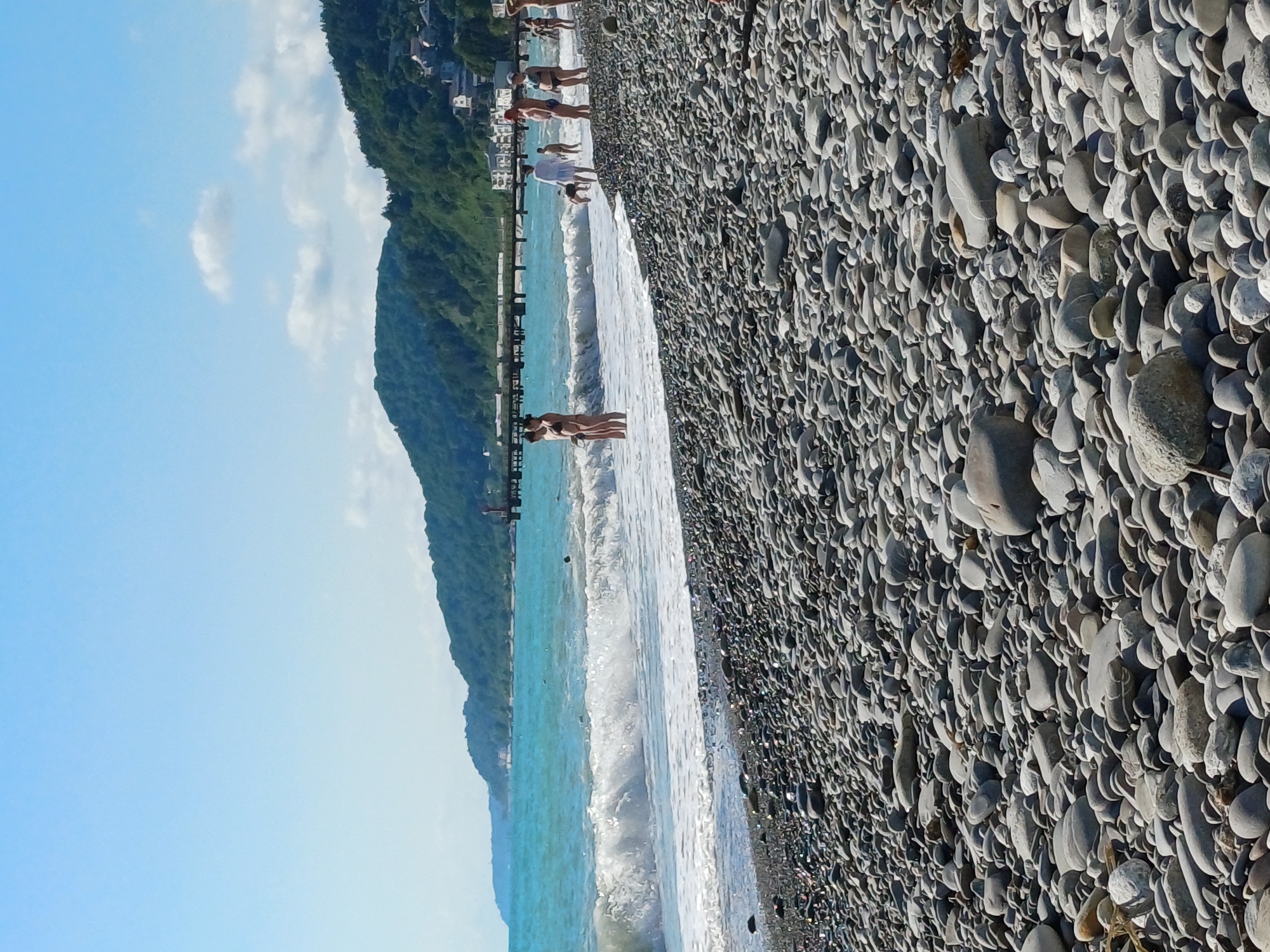 The image depicts a scenic view of a pebble beach in Sochi, Russia, under a clear blue sky with scattered white clouds. The foreground is dominated by a dense arrangement of smooth, grey pebbles of various sizes, reflecting the sunlight. In the middle ground, the turquoise waters of the Black Sea crash onto the shore with white foam. A long pier extends into the sea in the background.
Several people are visible on the beach and in the water. On the beach, individuals are seen standing, walking, and observing the sea. Some are dressed in swimwear, indicating a recreational atmosphere. In the water, a few figures are wading or swimming. A couple is standing close together near the water's edge. Further down the beach, a group of people are gathered, some standing and others partially in the water. A small dog is also present on the beach.
The background features a lush, green hillside covered with trees, with buildings scattered amongst the foliage. The overall scene conveys a sense of a popular tourist destination on a sunny day, likely during the summer season, given the attire of the people and the presence of swimmers. The location appears to be a typical coastal resort area in Sochi, known for its pebble beaches and mountainous backdrop.