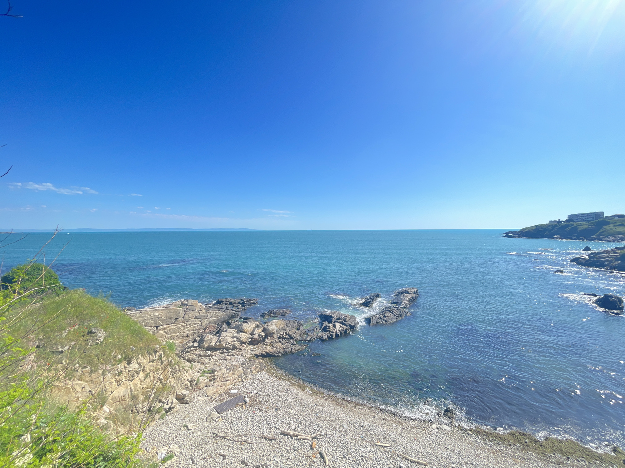 The image shows a coastal view in Sozopol, Bulgaria, on a clear, sunny day. The sky is a deep blue with a few wispy clouds near the horizon. The turquoise sea is mostly calm, with some visible wave action around rocky outcrops closer to the shore.
The foreground features a pebble beach and rocky cliffs covered with vegetation. In the distance, a headland with buildings is visible to the right, and another landmass is faintly discernible on the horizon.