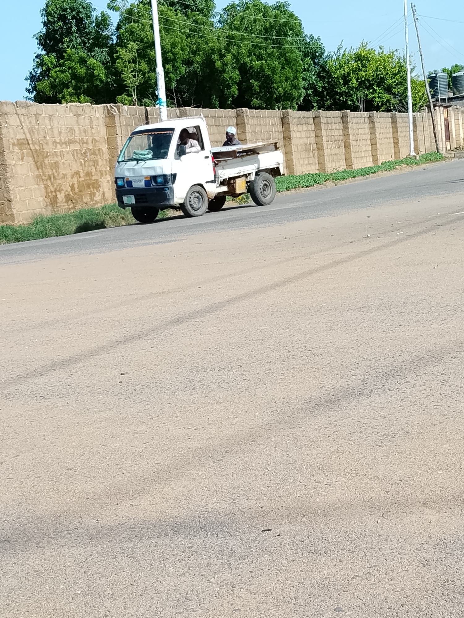 A white mini-truck is parked on the side of a road in Wauro Leggal, Nigeria. The truck has two people inside, a driver and a passenger. The truck's bed is empty. A brick wall runs along the side of the road, and there are trees and a utility pole in the background. The road appears to be paved and is dusty. The sky is clear and blue.