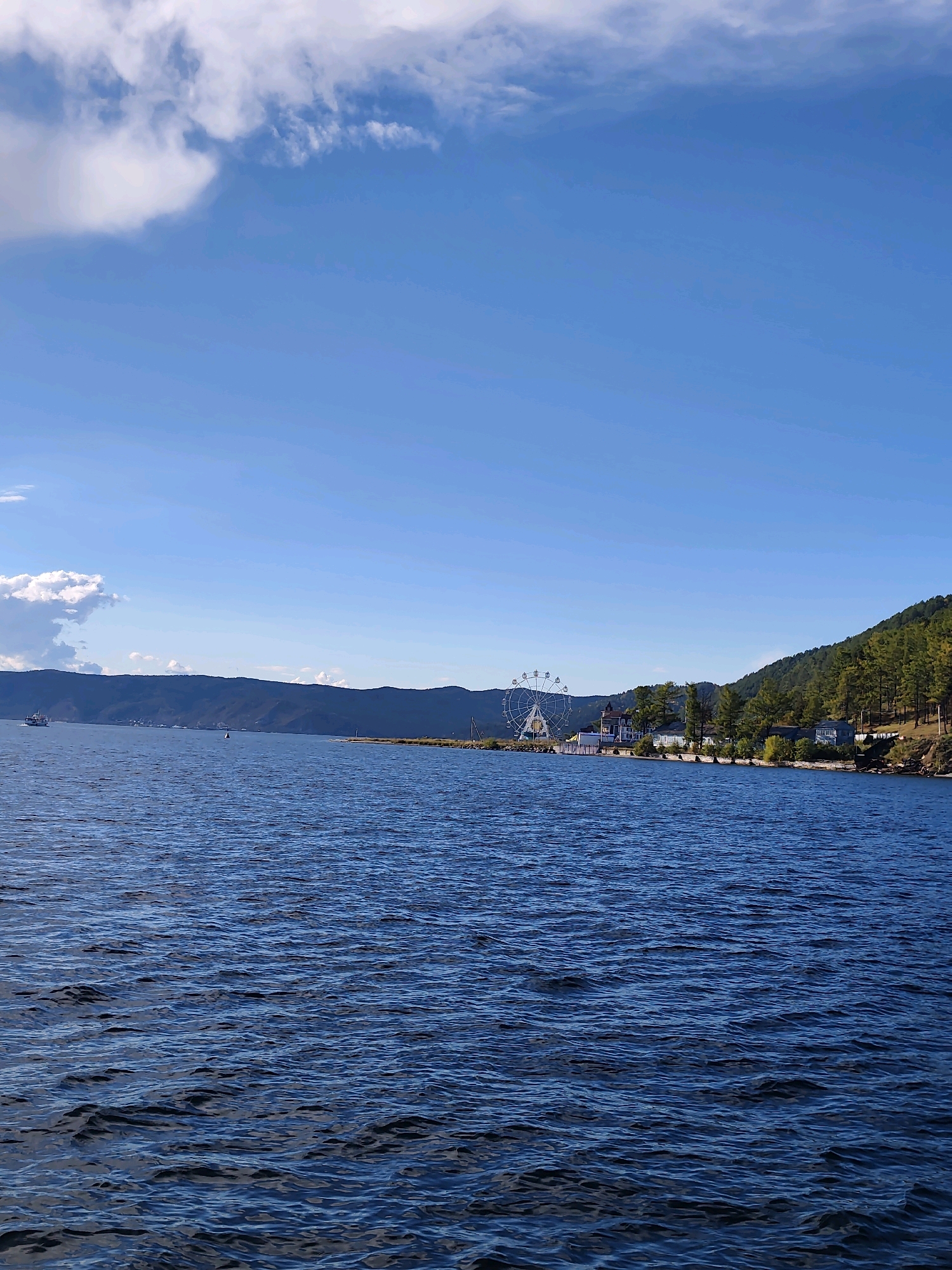 The image depicts a wide, calm body of water, likely Lake Baikal, under a bright blue sky with scattered white clouds. In the distance, a green, tree-covered mountain range forms the horizon. Along the shoreline, there is a Ferris wheel and several buildings, suggesting a developed area. A small white boat is visible on the left side of the water. The foreground is dominated by the deep blue water, with gentle ripples reflecting the sky. The scene appears to be a scenic view from a boat or the shore of Listvyanka, Russia, a popular tourist destination on the southern tip of Lake Baikal.