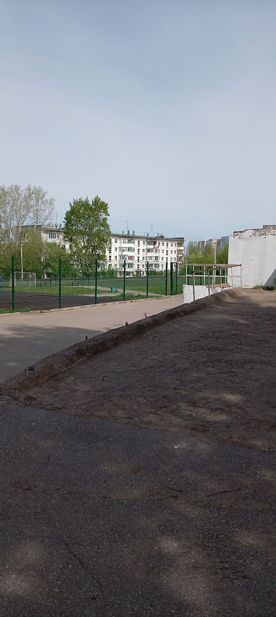 The image depicts a street scene in Chaikovsky, Russia. In the foreground, a dark, cracked asphalt surface occupies the lower portion of the frame. Above this, a slightly elevated sidewalk or curb runs horizontally. In the middle ground, there's a sports field enclosed by a green fence. Beyond the field, a white, multi-story apartment building is visible, punctuated by rows of windows and balconies. Scattered trees add a touch of greenery around the building and adjacent areas. To the right, a portion of a white concrete structure with a metal railing is present. The sky is overcast, contributing to a muted, somewhat gray overall tone. The scene appears devoid of people.