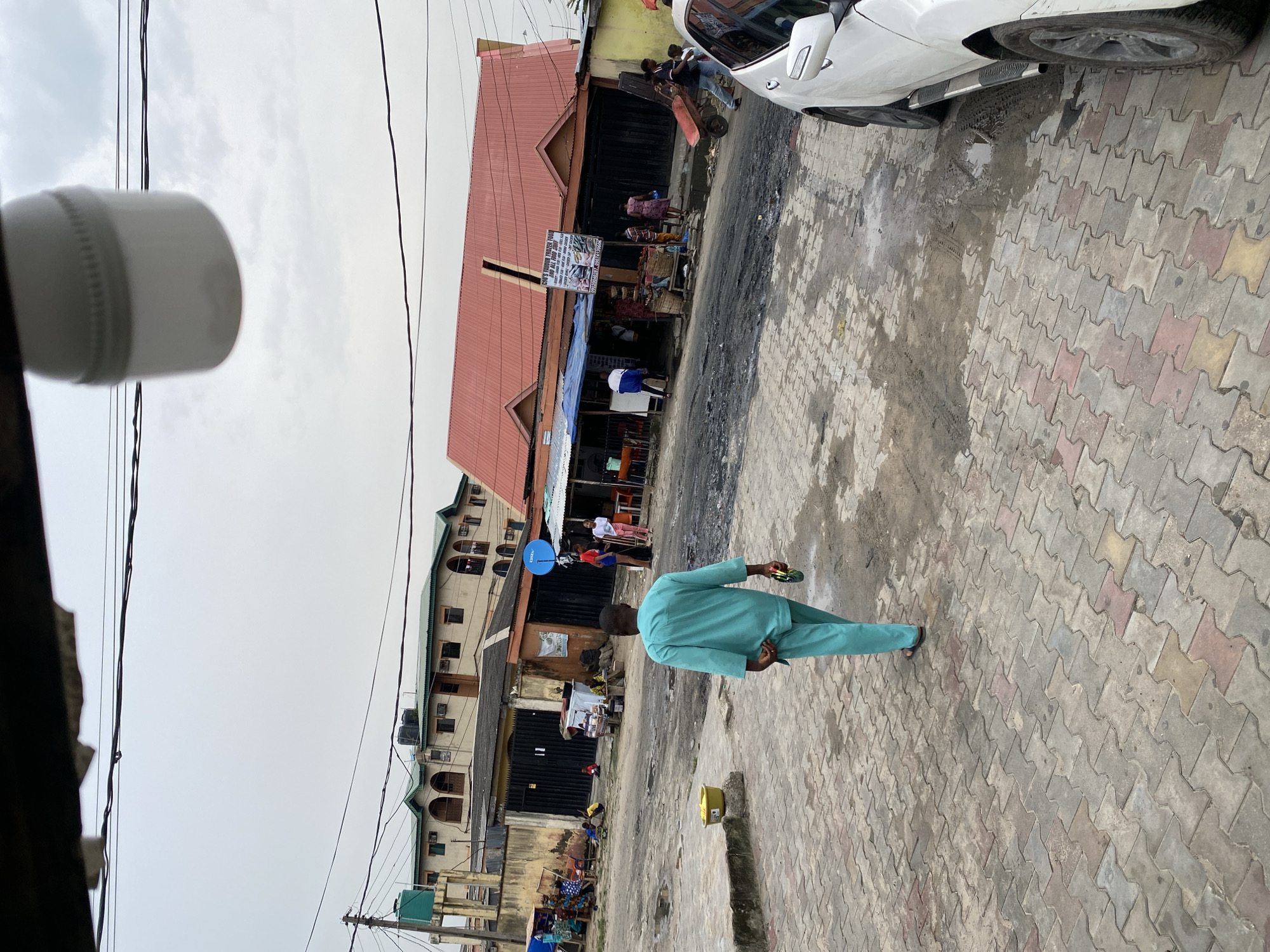 The image depicts a street in Ado, Nigeria. A man in a light blue suit walks away from the camera on a pavement made of interlocking pavers. Buildings are visible on both sides of the street, including a single-story structure with a red roof, appearing to be a storefront with people gathered around. Overhead power lines are prominent. A white SUV is partially visible on the right edge of the frame. A yellow container sits on the edge of the pavement near the man. Groups of people are seen further down the street near the buildings. The sky is overcast. A white object is visible in the upper left corner of the image.