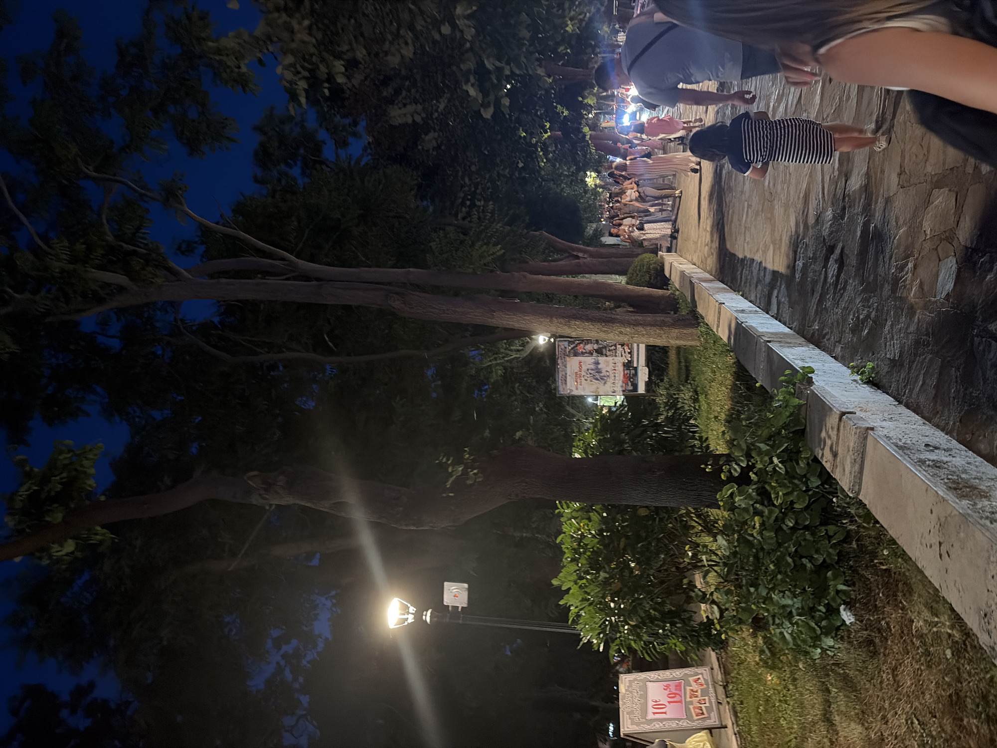 This is a nighttime photograph of a pedestrian walkway in Sozopol, Bulgaria. The walkway is paved with stone and lined with trees and a low concrete barrier. On the left side of the walkway, there is a grassy area with a small bush and a sign advertising retro photography. Further down the walkway, a lamppost illuminates the scene.
A group of people are walking along the path. In the foreground, a person is seen from behind, and a young girl in a striped dress is walking ahead of them. Further down, several adults are walking in the same direction. The sky is dark blue, indicating it is evening or night. The scene suggests a pleasant evening stroll along a promenade in the coastal town of Sozopol.