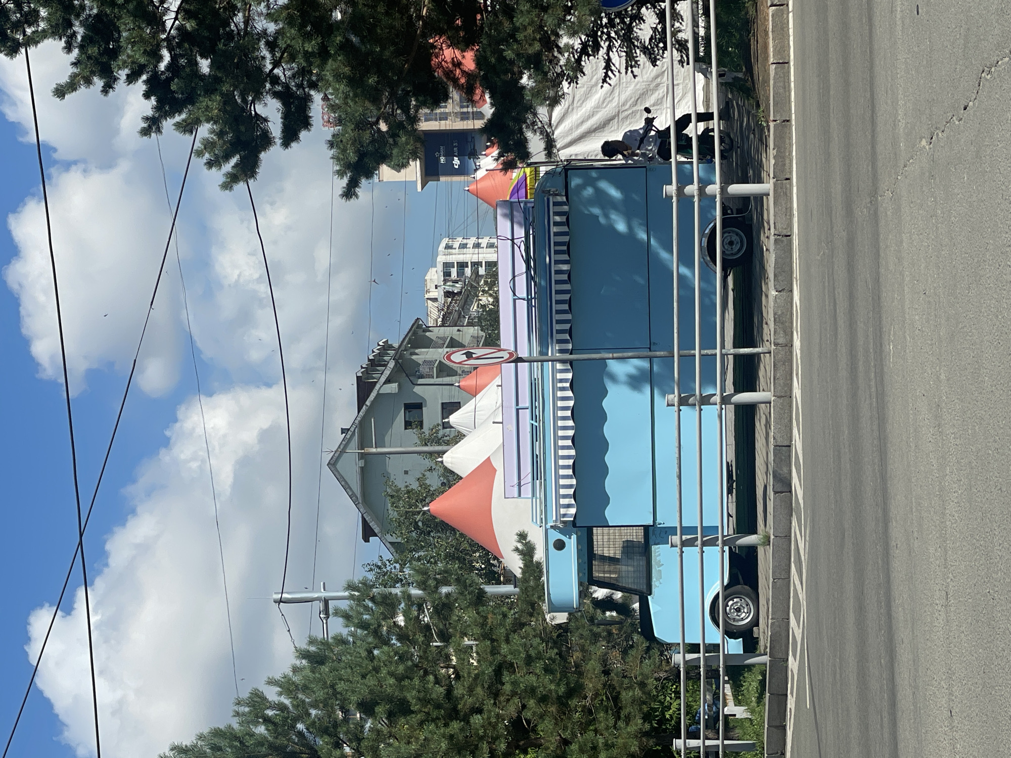 The image depicts a bright, sunny day in Ulaanbaatar, Mongolia, with a clear blue sky dotted with white, puffy clouds. The scene features a vibrant light blue food truck parked beside a metal railing on a paved street. The food truck has a striped awning and appears to be equipped for business. Behind the truck, several conical tents in red and white hues are visible, suggesting a possible event or market. A person is seen riding a green scooter to the right of the food truck, their back to the camera. The surrounding environment includes trees, buildings, and power lines, characteristic of an urban setting.