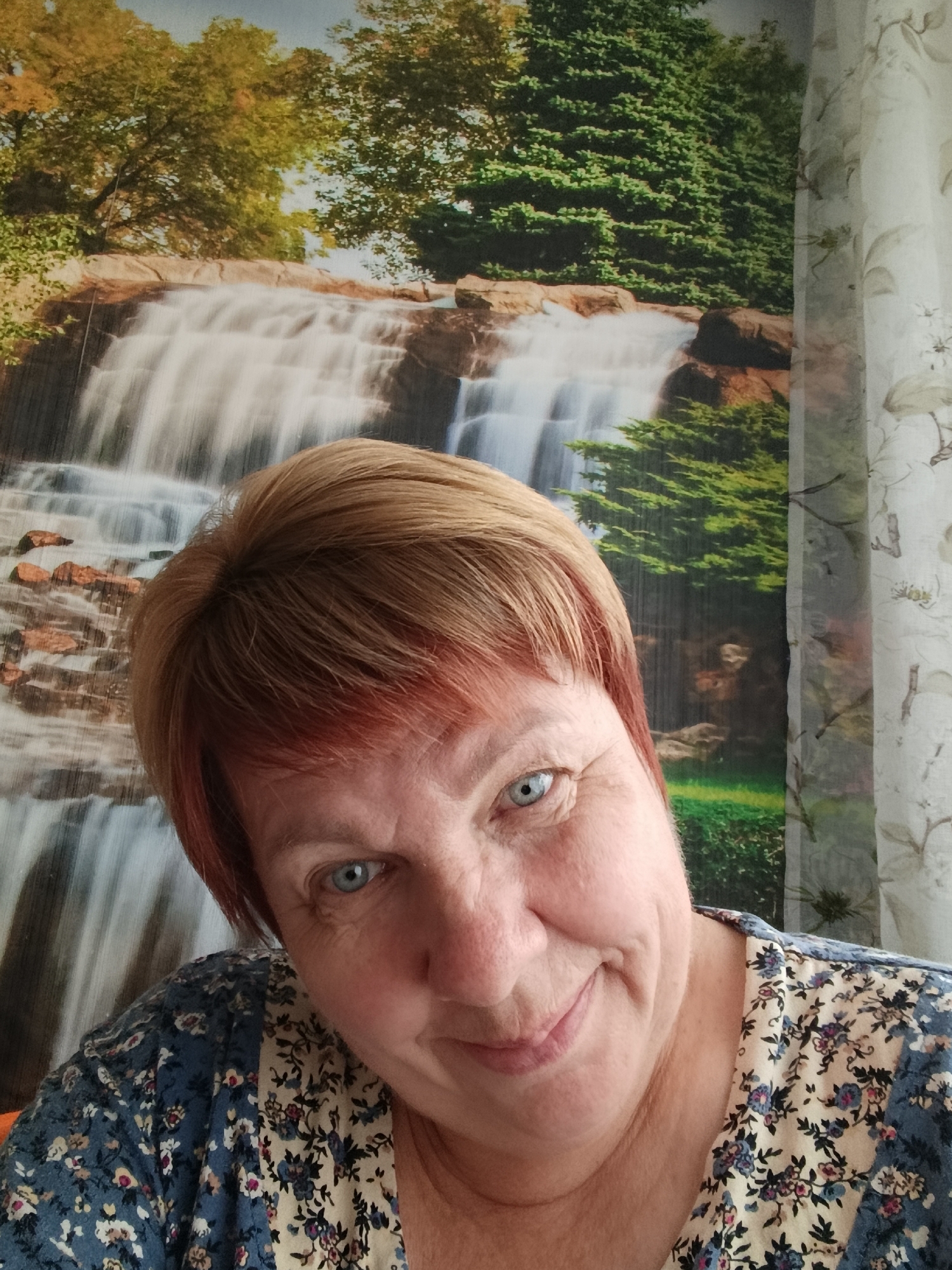 An elderly Caucasian woman with short, reddish-blonde hair and blue eyes is taking a selfie. She is wearing a patterned blouse. The background is a mural of a waterfall and lush green trees, and a curtain can be seen on the right side of the image. The image appears to have been taken indoors in a residential setting. There is no information in the image that can indicate a specific location within the city of Svietlahorsk, Belarus.