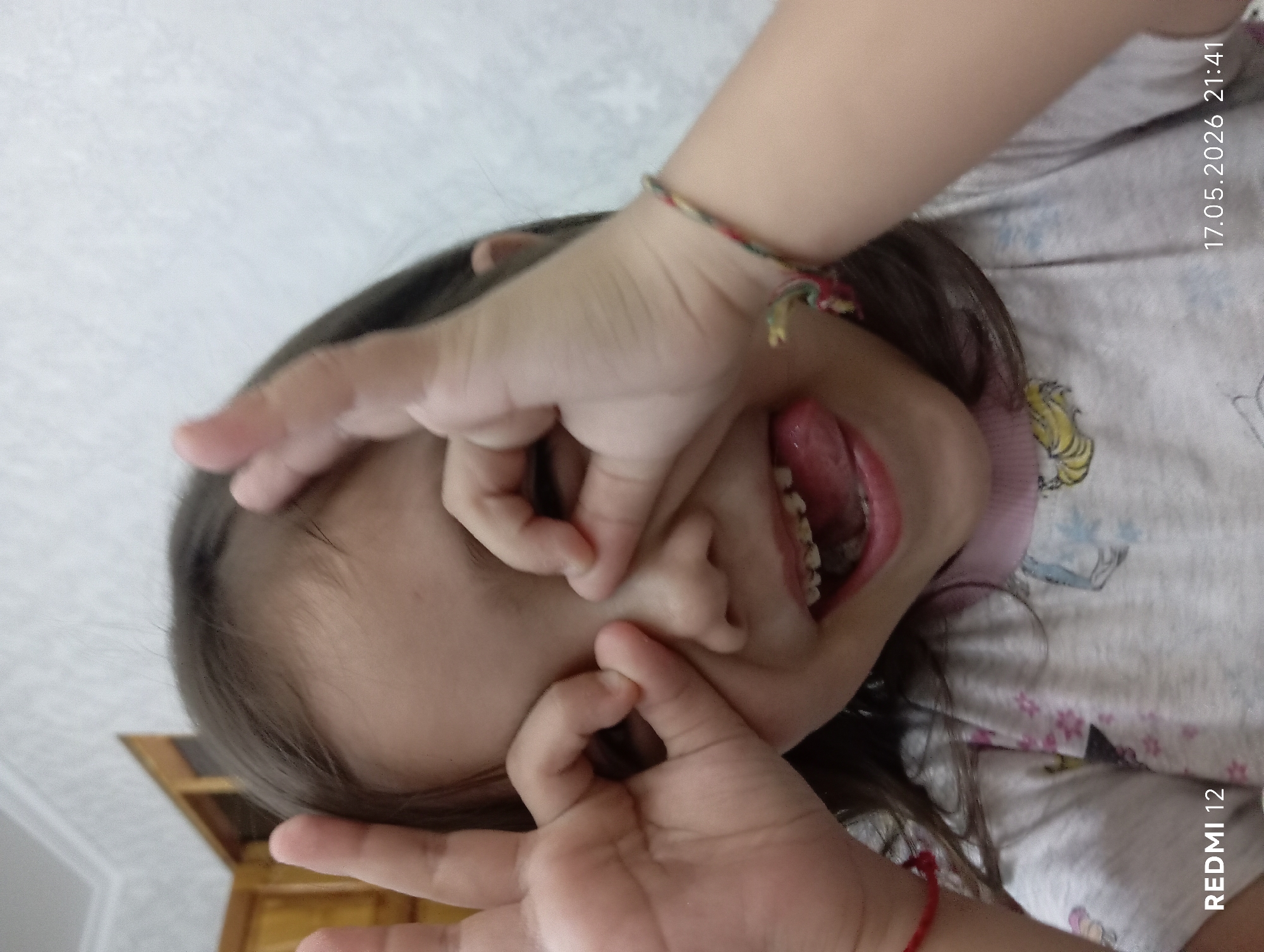 A child is holding their fingers in front of their face, distorting their nose. The background appears to be indoors, possibly a house. The timestamp in the top right corner indicates May 17, 2026, at 21:41. There is no visible evidence to confirm the location is Bekabad, Uzbekistan. The child is wearing a shirt with cartoon designs. A bracelet is on the child's wrist. The child has braces.