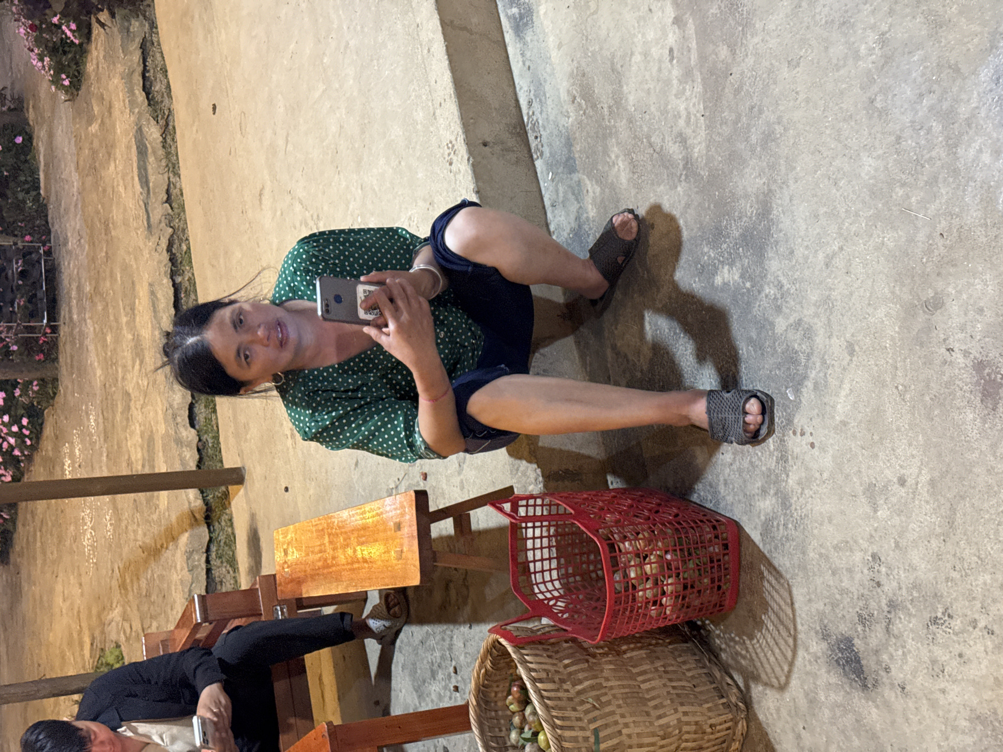 Captured from an elevated angle, the image showcases a woman seated on an outdoor concrete surface in Bắc Hà, Vietnam. She's engrossed in using a smartphone, clad in a green polka-dot top and denim shorts. Her footwear consists of grey sandals. Positioned adjacent to her are two baskets: one made of red plastic and the other of woven wicker. Both are filled with fruit. A second individual, partially visible, is seated at a wooden table with a bench, using a smartphone as well. The surroundings include outdoor seating and a view of stone structures and greenery, typical of the city.