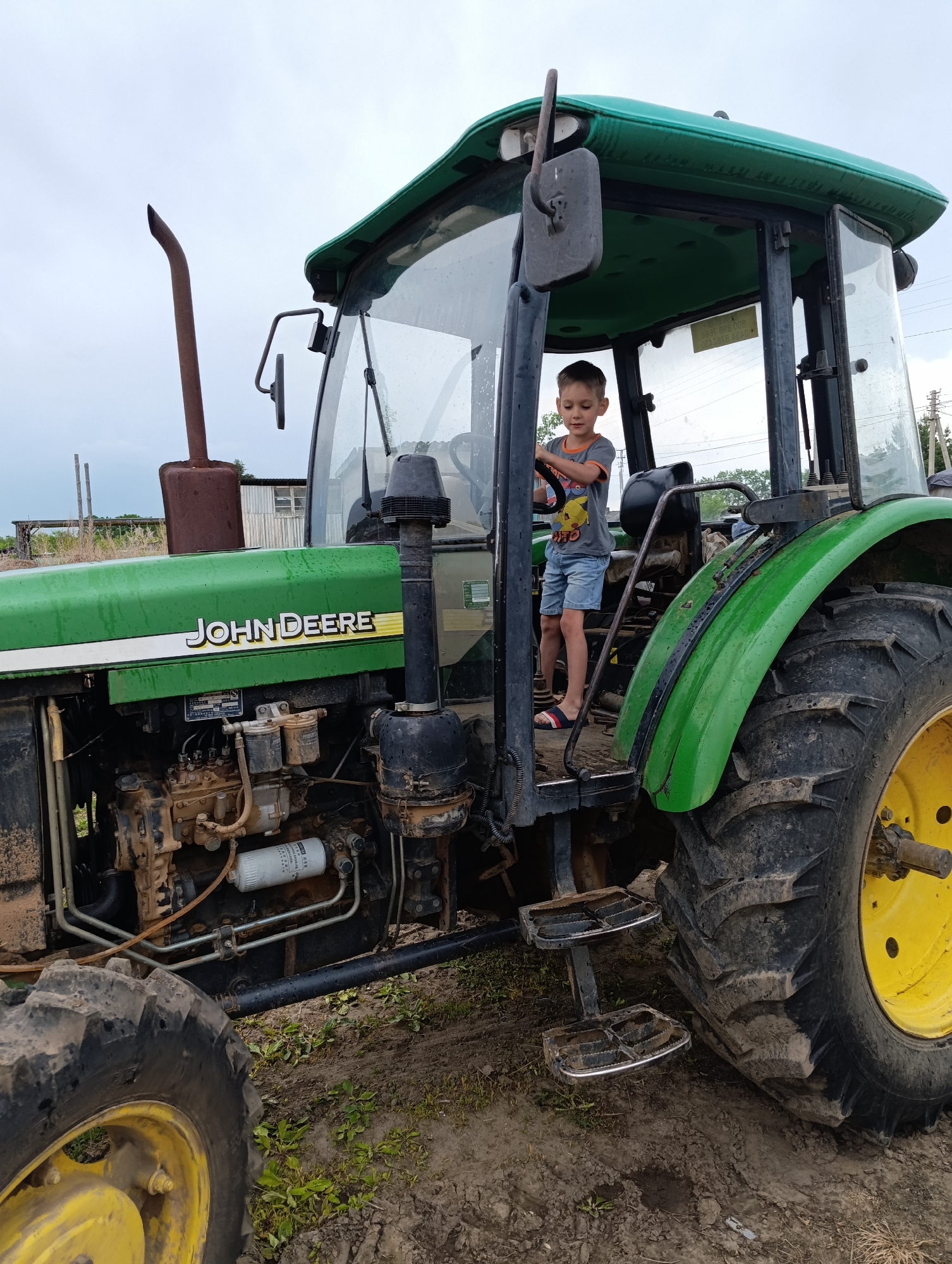 A boy stands in the driver's seat of a John Deere tractor in Dubovoye, Russia. The boy, who appears to be of primary school age, is wearing a gray t-shirt with a cartoon character and blue shorts. He is holding onto the steering wheel of the tractor. The tractor is green and yellow, with large, muddy tires. The cab of the tractor is enclosed with glass windows. The background shows a rural setting with some farm buildings and vegetation under a cloudy sky. The ground is muddy, indicating recent rain or wet conditions. The scene appears to be a casual moment of a child exploring a piece of farm machinery.