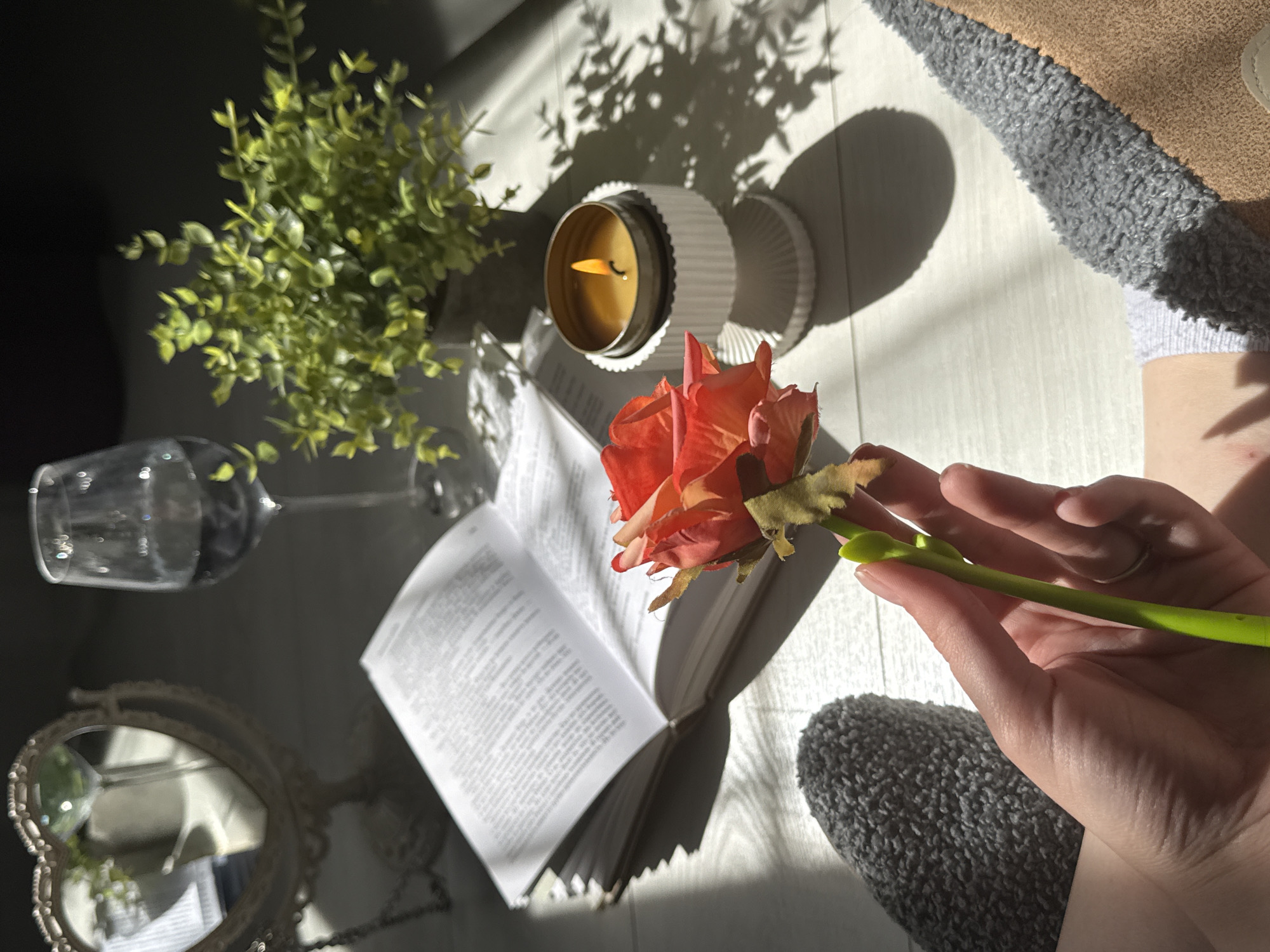 A hand, with a ring on the ring finger, is holding a single artificial rose with orange and red petals and a green stem. The rose is positioned in the foreground, slightly to the left of center. In the background, an open book with text is visible on a light-colored wooden surface. To the right of the book, a lit candle in a white decorative holder sits next to a small potted plant. A wine glass filled with dark liquid is also present. To the left of the book, a heart-shaped mirror is reflected in another mirror. The scene is illuminated by sunlight casting shadows and creating a warm atmosphere. A grey, fuzzy sock is visible on the left foot, and a tan slipper is on the right foot, suggesting the person is relaxing at home.