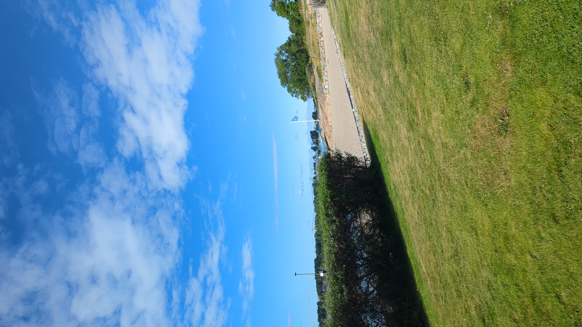 The image shows a sunny day with a clear blue sky filled with scattered white clouds. In the foreground, a lush green lawn slopes gently downwards. A dark green hedge runs along the left side of the frame, separating the lawn from a sandy beach area. The beach is bordered by a low stone wall and a paved path. Further in the distance, a tall flagpole with a blue flag stands against the backdrop of trees and buildings. The water is visible behind the hedge, and the overall scene suggests a peaceful outdoor location, likely a park or recreational area near the coast. The location is Larkollen, Norway.