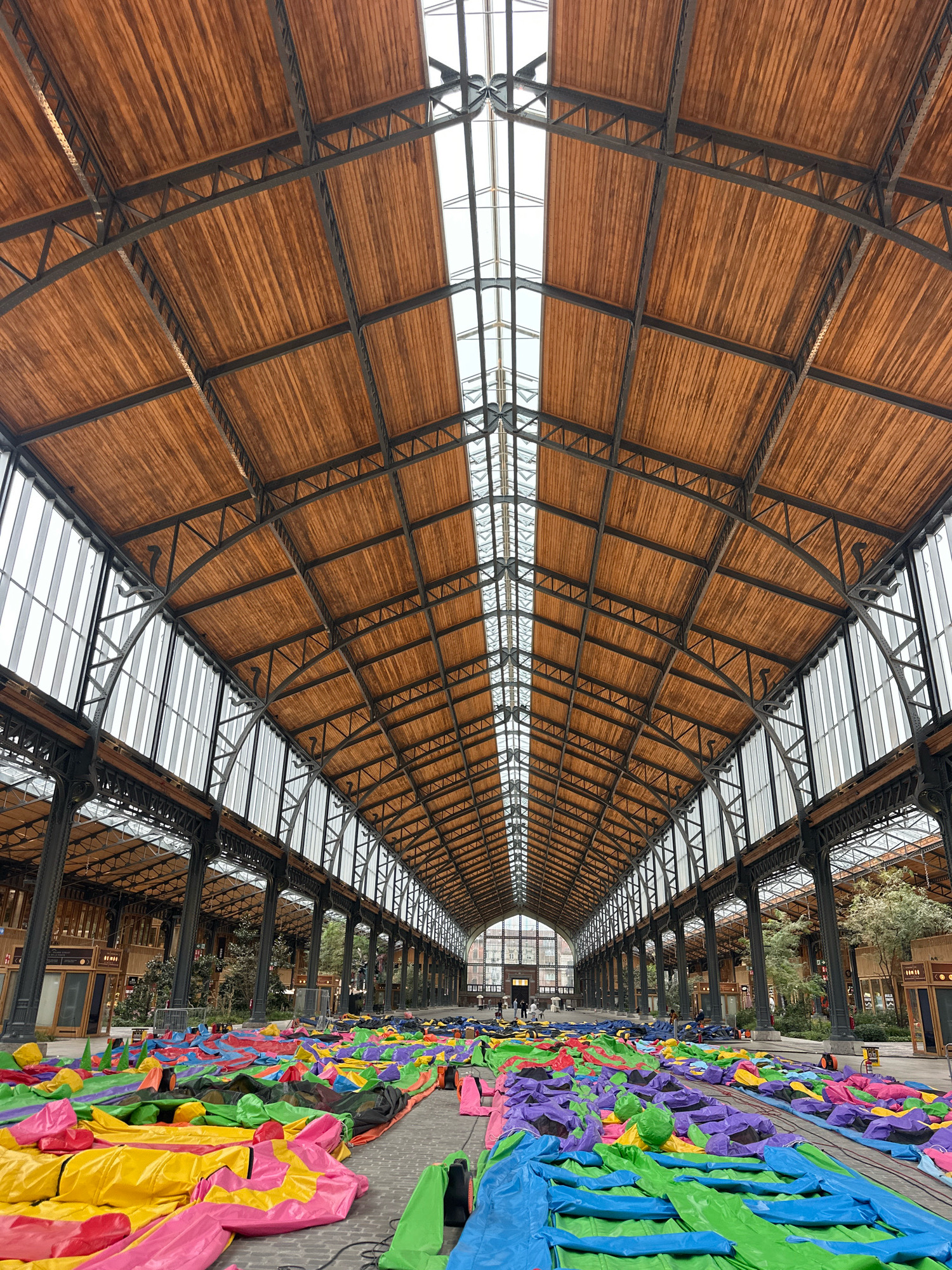 The image shows the interior of a large, historic building with a vaulted wooden ceiling supported by a dark metal frame. Large windows line the sides of the building, letting in natural light. The floor is covered with a vast expanse of deflated, brightly colored inflatable structures in shades of pink, yellow, green, blue, and purple. Some of these inflatables appear to be arranged in a pattern, while others are jumbled together. In the background, a few people can be seen walking, and there are trees and what appear to be market stalls or shop fronts along the sides of the building. The scene suggests preparation for an event or activity involving these inflatable structures. The architectural style of the building, particularly the ornate metalwork and vaulted ceiling, is characteristic of historic European train stations or market halls. This specific location is the Gare Maritime (Maritime Station) in Tour & Taxis, Brussels, Belgium, a former railway goods station that has been redeveloped into a public space.