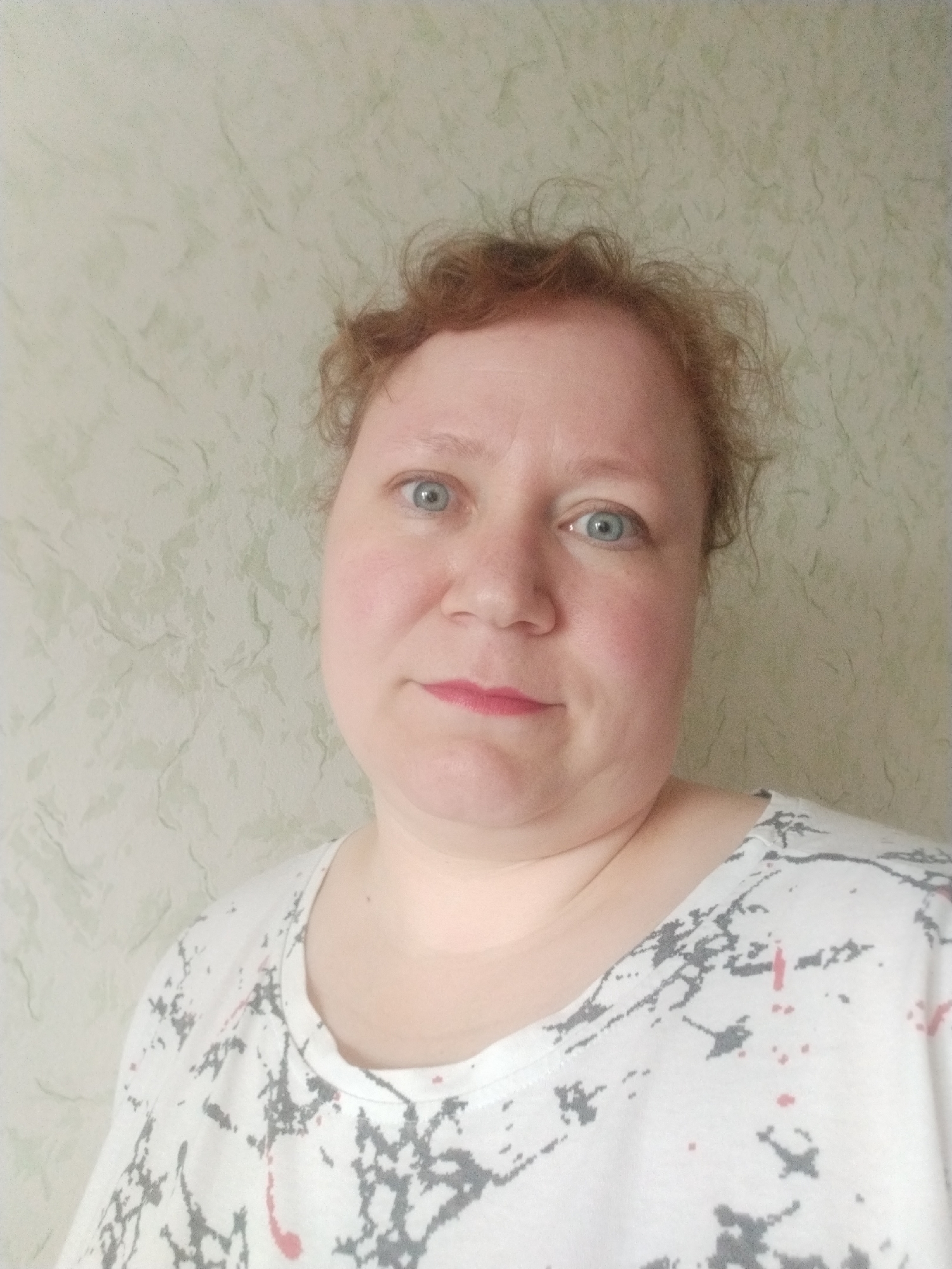 A woman with fair skin, light blue eyes, and reddish-blonde hair is facing the camera. She is wearing a white T-shirt with a gray and pink speckled pattern. The background is a light green wall with a subtle, marbled texture. The picture was taken indoors. There is no information about the location being in Chaikovsky, Russia.