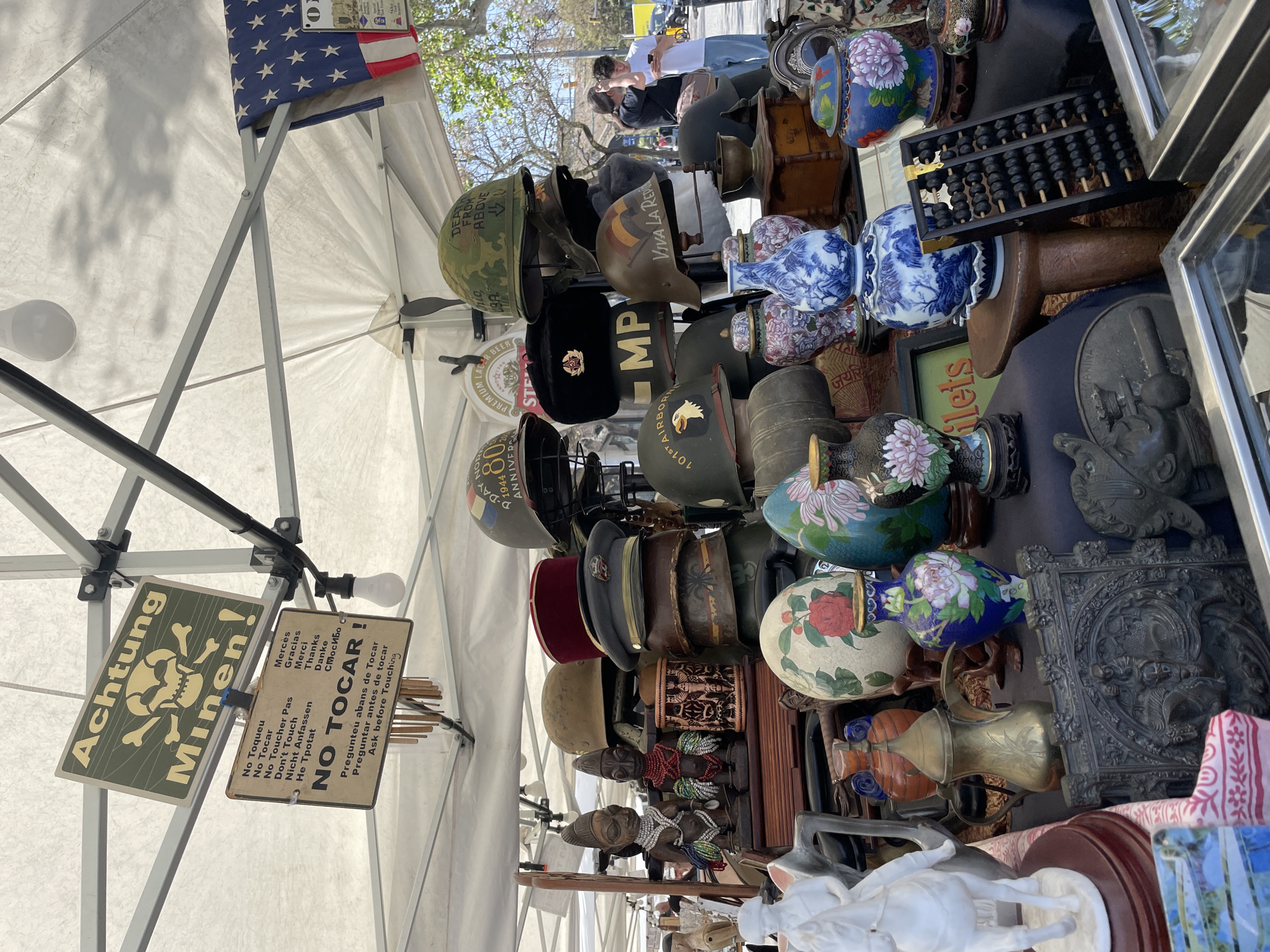 This image shows an outdoor display of vintage items likely at a flea market or antique fair. The items include military helmets, decorative vases, figurines, and other miscellaneous objects such as an abacus and small wooden figures. A sign reading "Achtung Minen!" is visible, along with a "No Tocar" sign in multiple languages. People are visible in the background, suggesting a busy marketplace setting. The presence of signs in Spanish and Catalan, along with the phrase "Pregunteu abans de Tocar," indicates that this market is located in Barcelona, Spain.