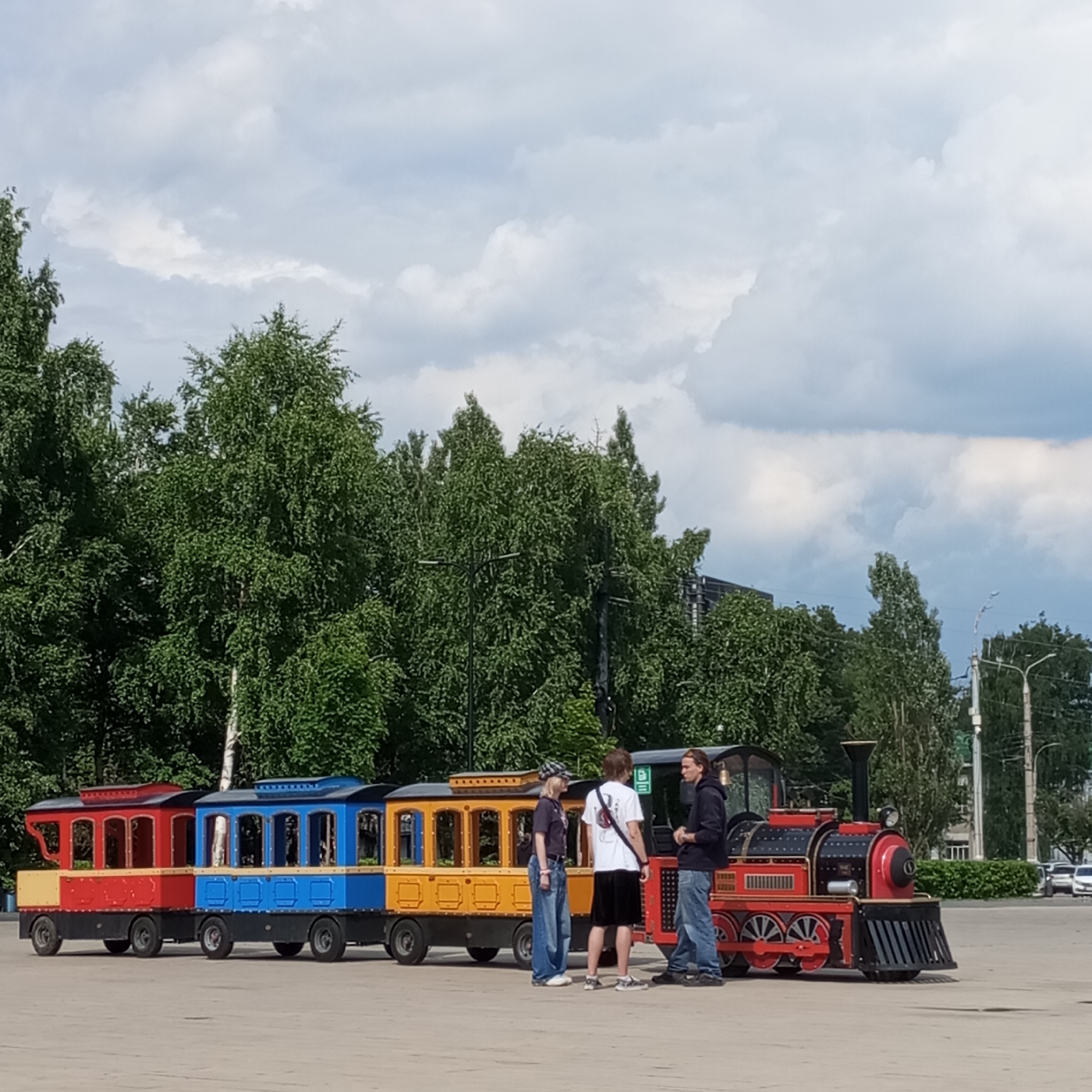 In Izhevsk, Russia, on a cloudy day, a miniature train with a red engine and colorful cars (red, blue, yellow) is parked on a paved surface. Three young individuals are standing near the train. One, a male with long hair, is on the far right, wearing a dark hoodie and jeans. To his left, a male in a white t-shirt and black shorts is facing the other two. On the left, a female wearing a dark shirt, jeans, and a patterned cap is facing the male in the white t-shirt. The train appears to be a recreational attraction. Behind the train, a line of lush green trees forms the background, with some buildings and streetlights visible in the distance.