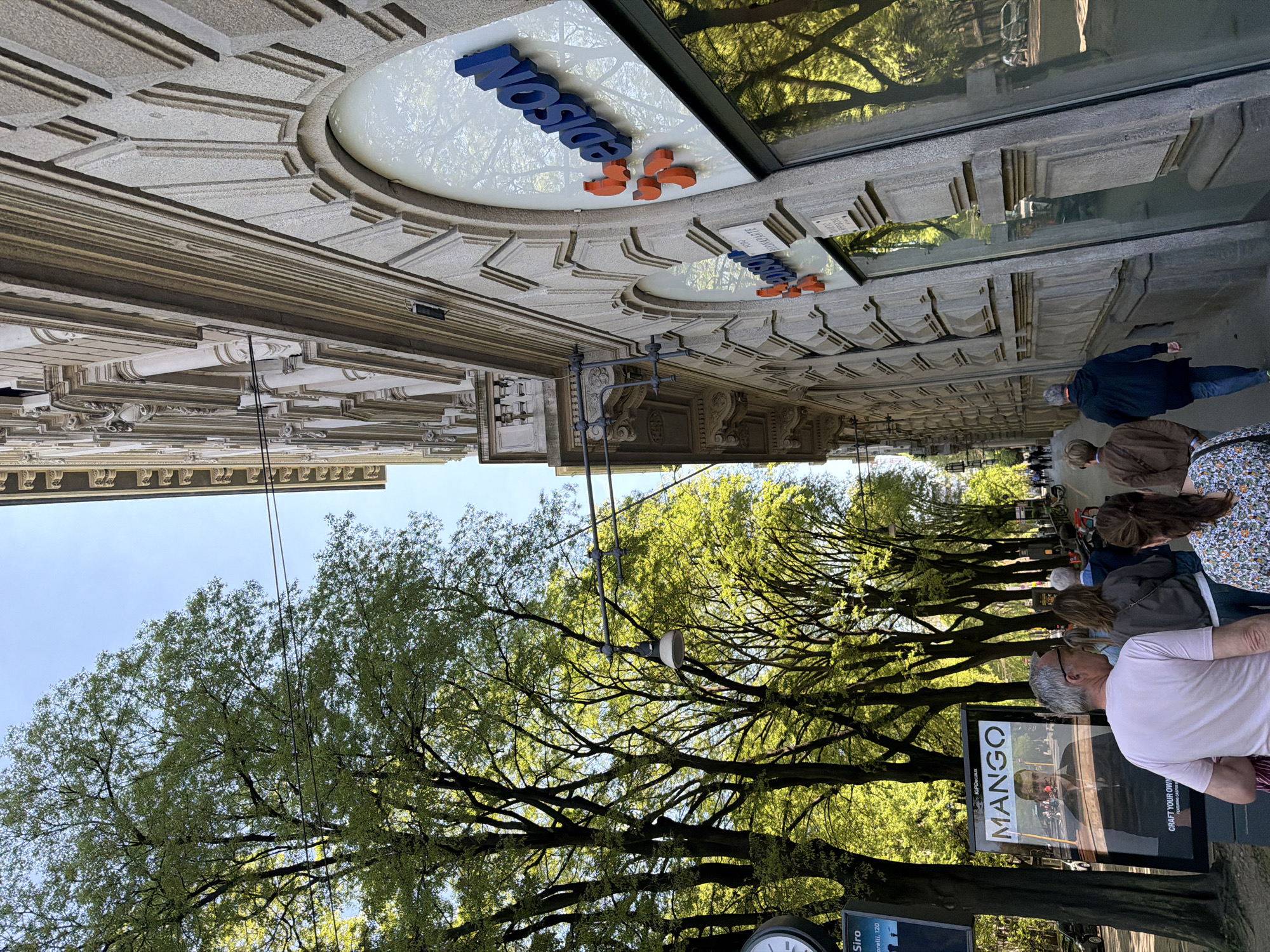 This image depicts a street scene in Milan, Italy, with a row of trees lining the pedestrian walkway. Several people are walking along the street. A "Mango" advertising poster is visible on the right. The building on the left bears the name "Edison" in blue and orange letters above a commercial space. The image is oriented upside down.