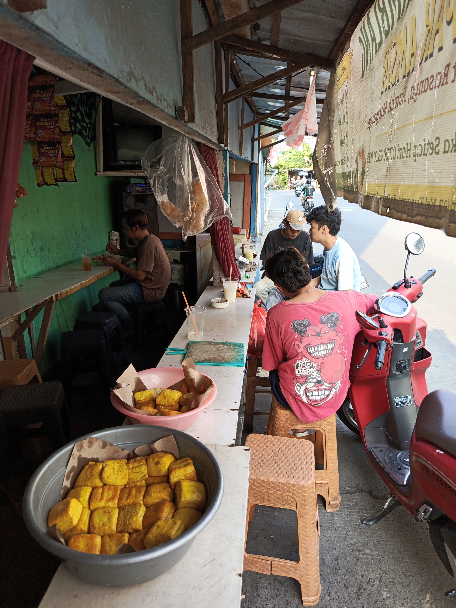 A street-side food stall in Tangerang, Indonesia, displays fried tofu in a large gray bowl and a smaller pink bowl, with one person eating at a green-walled counter and three young men seated on plastic stools near a red scooter. The stall has a wooden roof supported by beams, with a white banner advertising food hanging in the background. Bags of snacks are suspended from the ceiling. The ground is paved with concrete. A red scooter is parked to the right of the frame. The time of day appears to be daytime, with sunlight casting shadows.