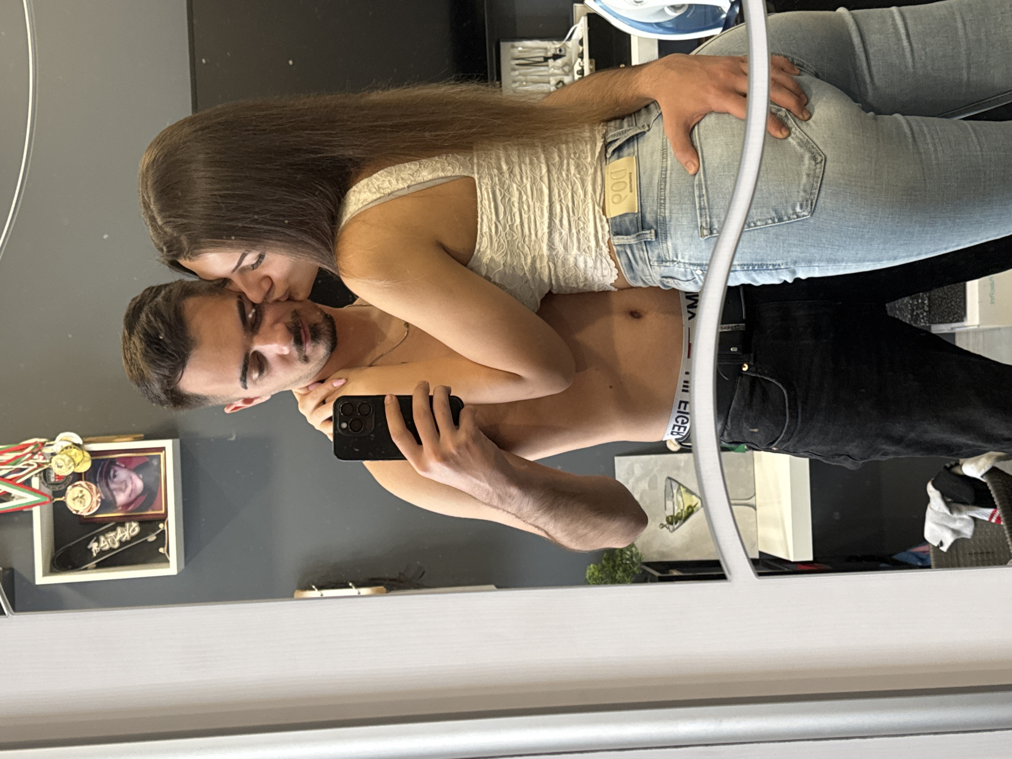 A man and a woman are visible in a mirror selfie, with the woman kissing the man's forehead. The man is shirtless with black jeans, holding a phone in his hand. The woman has long brown hair, wearing a white top and light blue jeans. Behind them, in the reflection, there is a wall with decorations, including what appears to be medals, a framed picture, and a skateboard. There is also an ironing board and various items on a table. The environment is somewhat cluttered. It is impossible to determine the specific location in Sofia, Bulgaria from the given image.