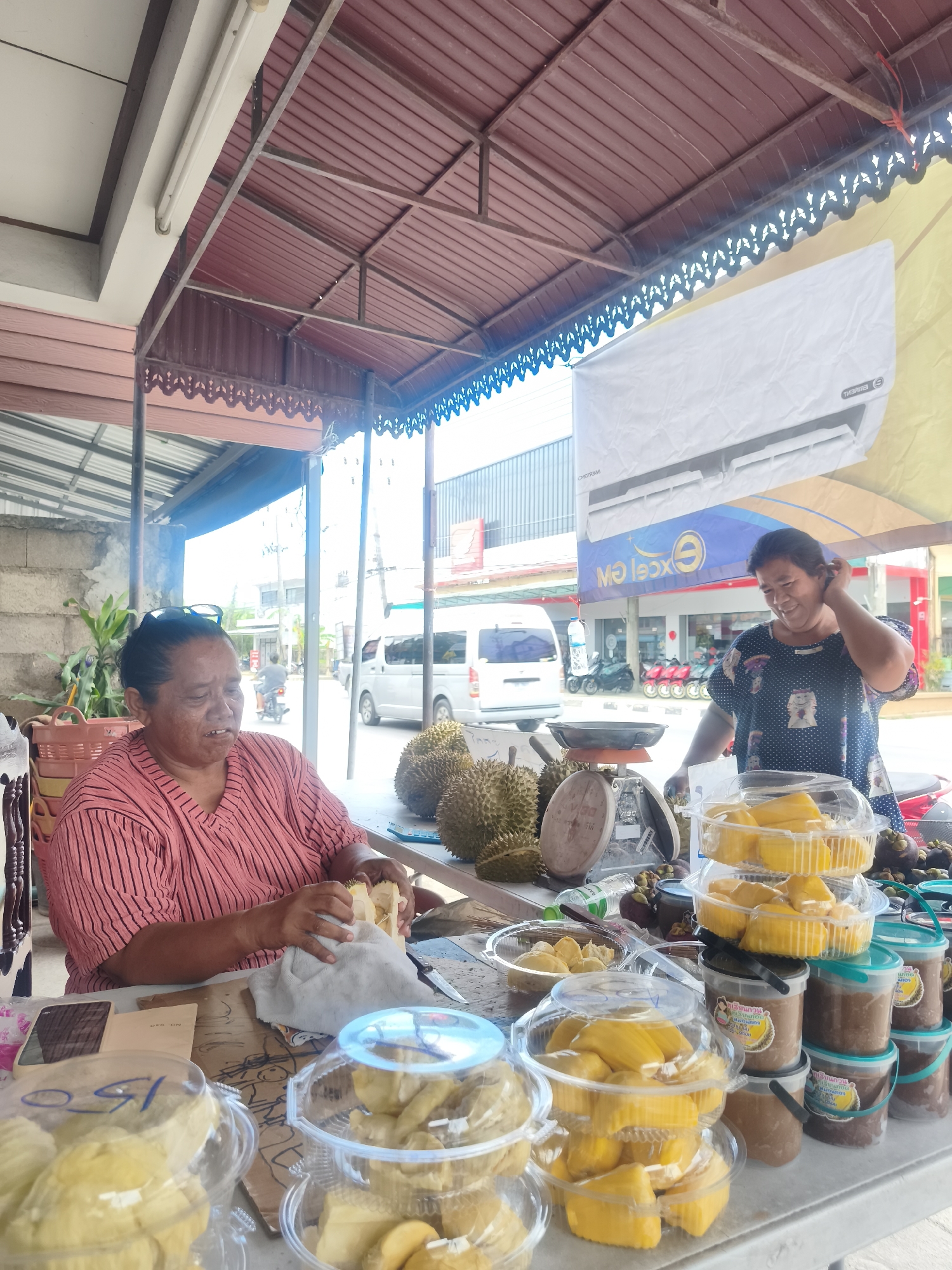 Two women are seen at an outdoor fruit stand. One woman, on the left, is wearing a pink and black striped shirt and is seated at a table, in the process of peeling a durian. She has sunglasses on the top of her head. In front of her are containers of cut jackfruit, a basket, and a mobile phone. Two whole durians are on the table to her right.
The second woman, on the right, is wearing a dark blue dress with white polka dots and is standing behind the table. She is smiling and touching her hair. In front of her are several containers of cut jackfruit, as well as tubs of what appears to be a paste or sauce. A scale is visible on the table.
The fruit stand is covered by a corrugated metal roof supported by metal poles. In the background, there is a street with vehicles, including vans and motorcycles. Buildings are visible on the other side of the street. A large white sheet with an air conditioning unit advertisement is hanging from the roof. The scene appears to be in a tropical climate, given the fruits displayed and the outdoor setting.