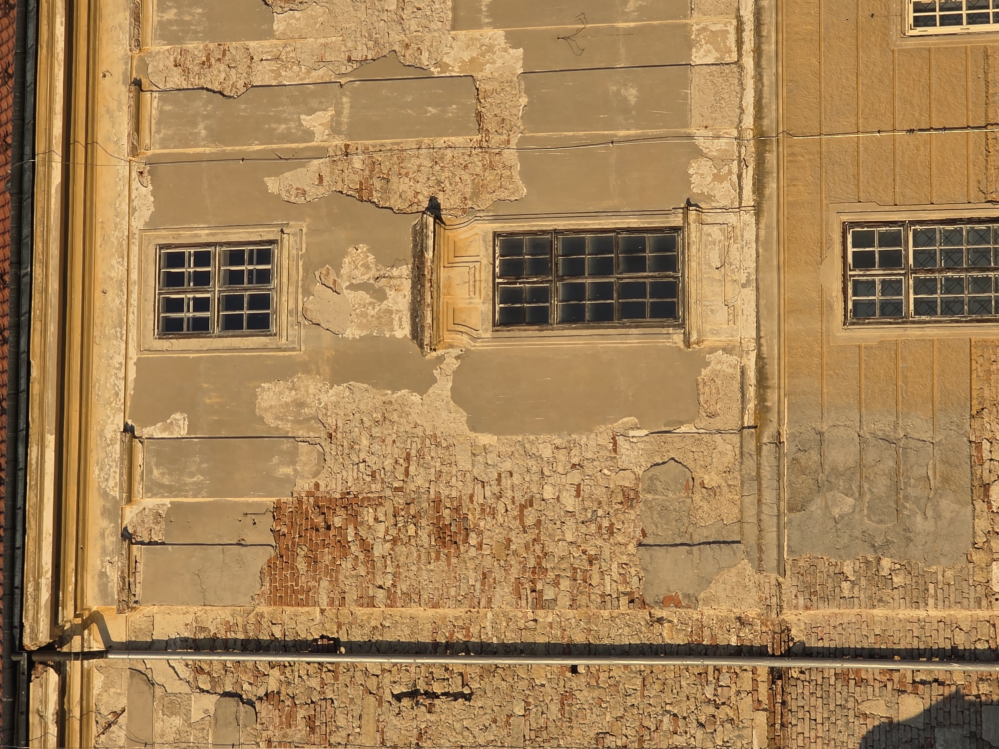 The image shows a close-up of the facade of an old, weathered building. The wall is constructed of exposed brick on the left side, with sections of crumbling plaster revealing the brickwork underneath on the right. The plaster is a light tan color and shows significant signs of decay, with large patches missing.
There are three windows visible on the facade. The topmost window is small and rectangular with a dark frame and multiple panes. It is set within a lighter colored frame with decorative architectural details. Below this is a larger, taller window with a similar dark frame and many panes, also surrounded by an ornate, lighter-colored frame. The bottom visible window is also rectangular with a dark frame and panes, set within a section of the wall that has horizontal decorative lines. A metal downspout runs vertically along the left side of the building, casting a shadow. The roofline is visible at the very top, with dark shingles. The sunlight appears to be warm and golden, suggesting it is either early morning or late afternoon. The overall impression is one of age and disrepair, with a stark contrast between the exposed brick and the decaying plaster.