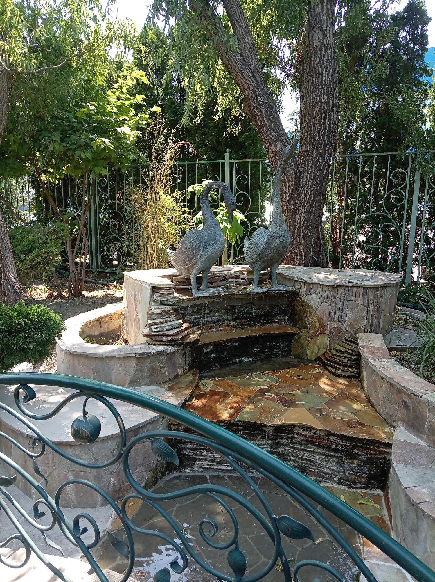The image shows a decorative tiered fountain with two goose sculptures in a garden setting, likely in Inozemtsevo, Russia. The fountain is constructed from stone and slate, with water flowing down the levels. The goose sculptures are made of a dark gray material and are positioned on the upper tier of the fountain. Behind the fountain, a large tree trunk is visible, and further back, there is a green metal fence with decorative scrollwork. Lush green foliage and trees surround the fountain, and sunlight filters through the leaves. In the foreground, a dark green metal railing with ornate scroll and leaf designs is present. The ground around the fountain is paved with irregular stone tiles. The scene appears to be a well-maintained outdoor space.