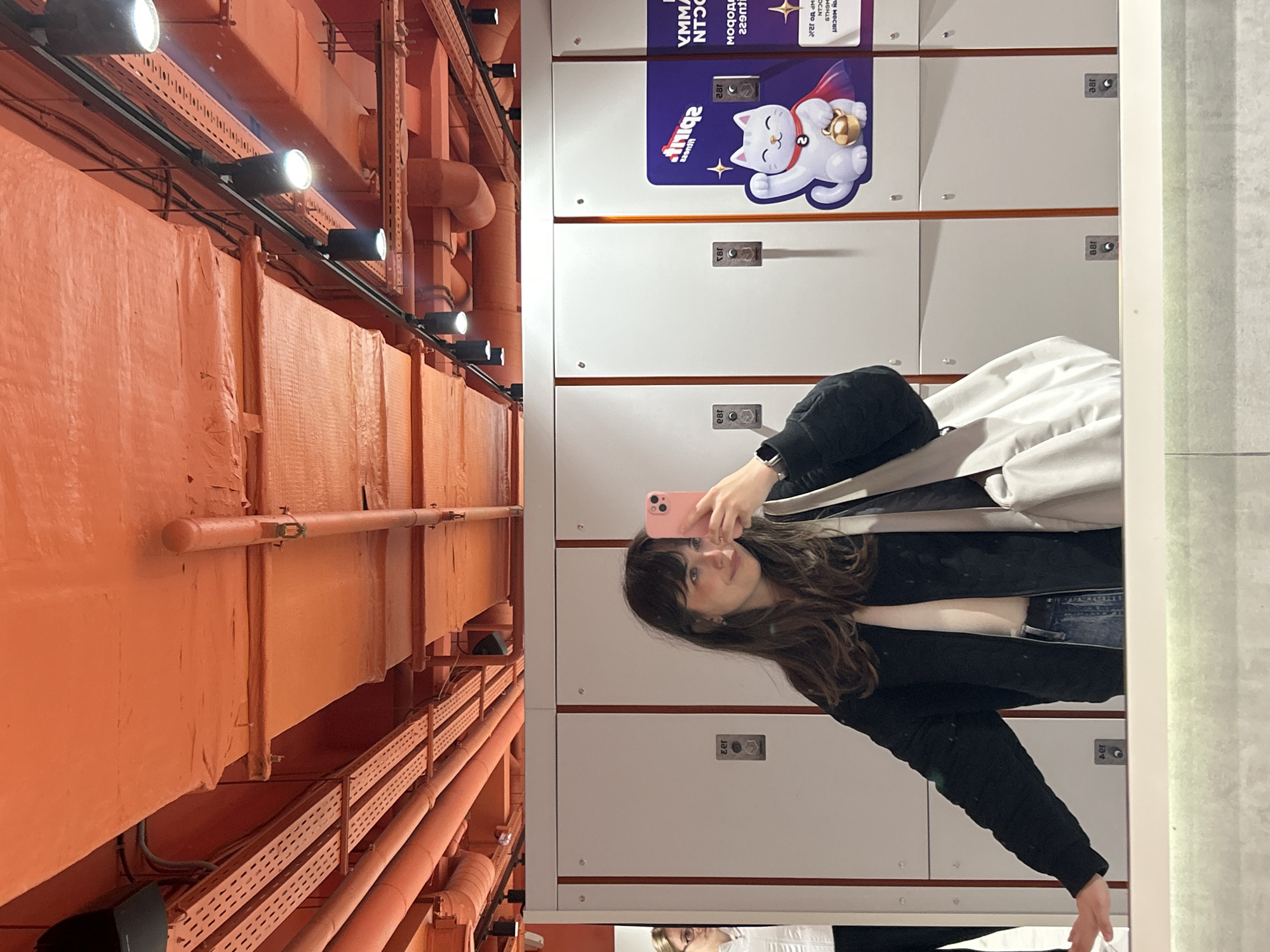 The image shows a woman taking a selfie in a mirror in what appears to be a locker room. The lockers are white with orange trim and numbered with "181" and "182". A sticker with a cartoon cat and Russian text is attached to the top lockers. The room has orange ceilings with pipes and lights running along them. It is likely to be located in Saint Petersburg, Russia due to the Russian text and cultural imagery.