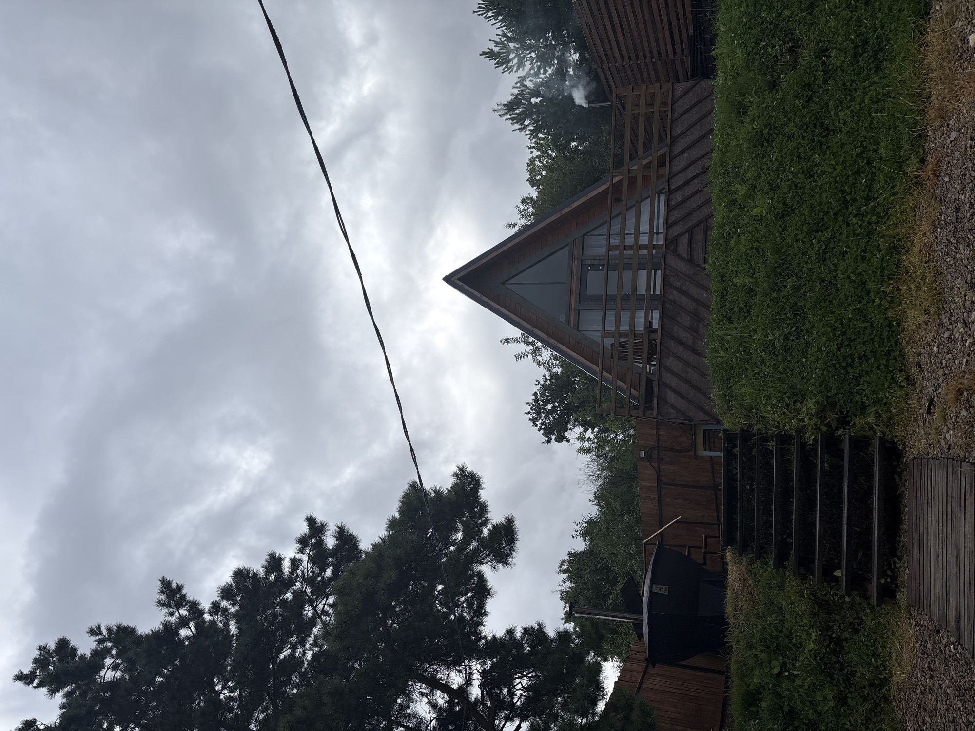 The image depicts a triangular wooden house with a large glass facade and a wooden deck in front, situated amidst a forest setting. To the left of the house, there is a dark, conical structure, possibly a hot tub or a sauna, with a wooden fence surrounding the property. A set of wooden stairs leads up to the deck. The sky is overcast with clouds. In the background, there are more trees and a smaller wooden structure. Smoke is seen coming out of a chimney to the right of the house. The scene is likely in the city of Chaikovsky, Russia, given the mention of the location. The overall atmosphere is serene and rustic.