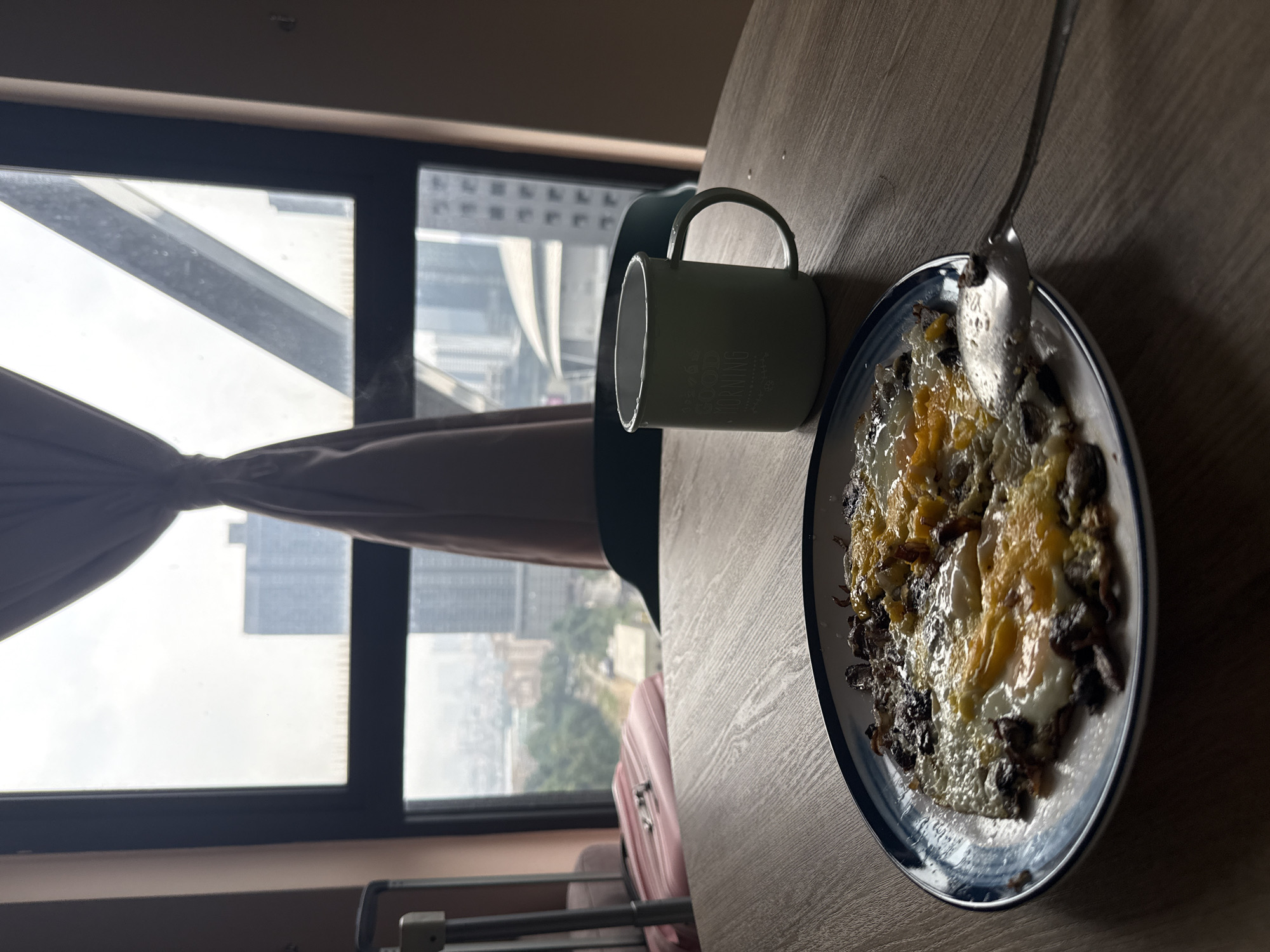A close-up, eye-level shot captures a table with a breakfast of eggs and meat, alongside a mug, placed in front of a window overlooking a cityscape. The eggs are fried with visible yolks and whites, mixed with dark pieces of meat. A silver spoon rests on the edge of the plate. The mug, a light green enamel cup, is positioned to the right of the plate and has "GOOD MORNING" printed on it in white text. Steam is rising from the mug, indicating a hot beverage.
The background reveals a large window with a view of Kuala Lumpur, Malaysia. Tall buildings and structures are visible through the glass, with some trees and greenery scattered throughout the urban landscape. A light brown curtain is partially drawn to the left of the window. To the left of the window, a pink suitcase with a silver handle is partially visible, and what appears to be a rolling suitcase or luggage stand is to its left. The overall lighting suggests it is daytime, likely morning, with soft natural light filtering through the window. The table surface is made of wood with a distinct grain pattern. The image appears to be taken from an interior location, possibly a hotel room or apartment, given the view and the luggage.