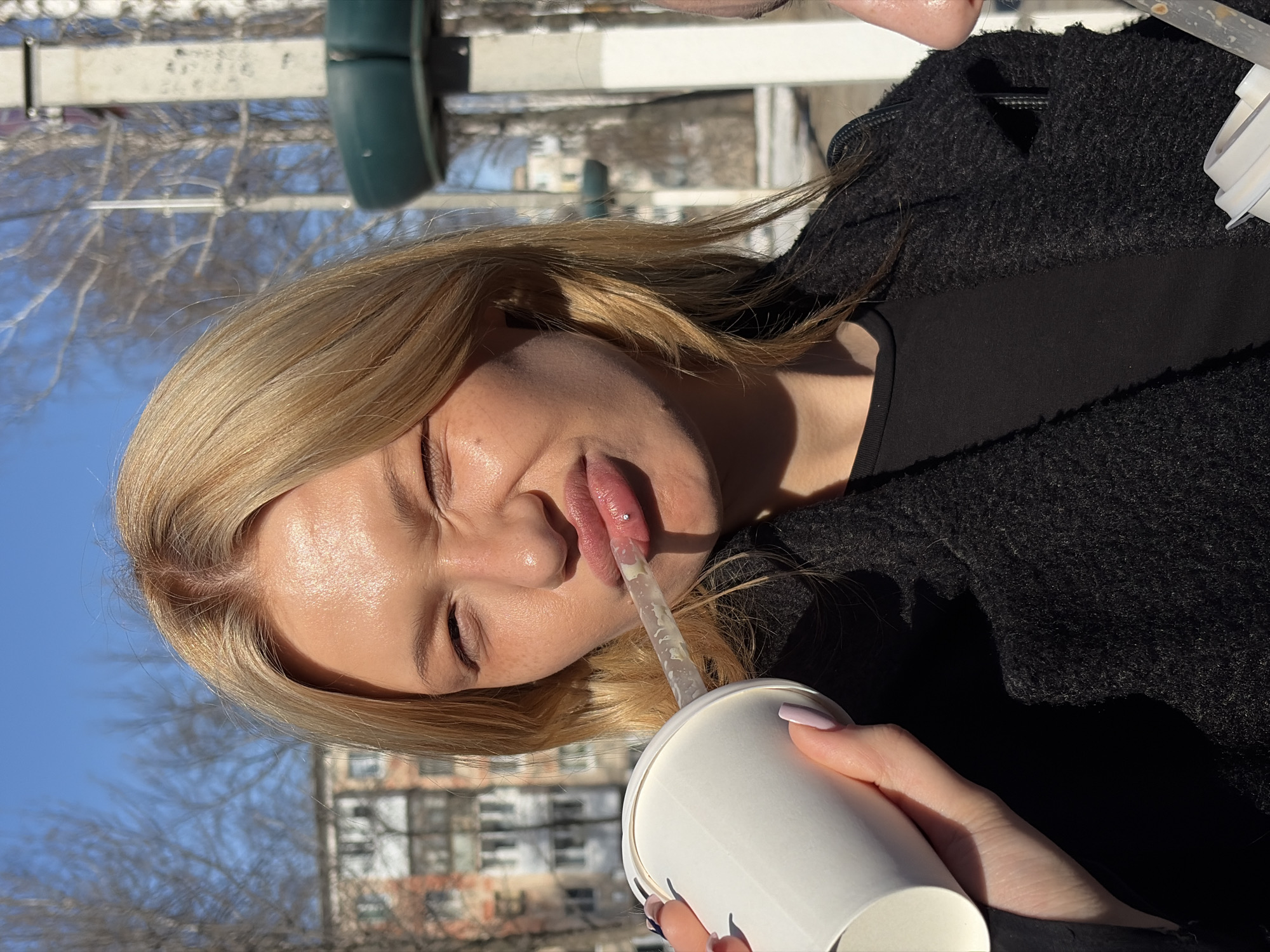 A young, fair-skinned woman with blonde hair is outdoors, likely in the city of Chaikovsky, Russia, based on background buildings resembling typical Russian urban architecture. She is drinking from a white paper cup with a straw. The woman is wearing a dark, possibly black, textured jacket or coat over a black top. The setting appears to be a sunny day, with bare trees in the background and building facades that suggest an urban environment.