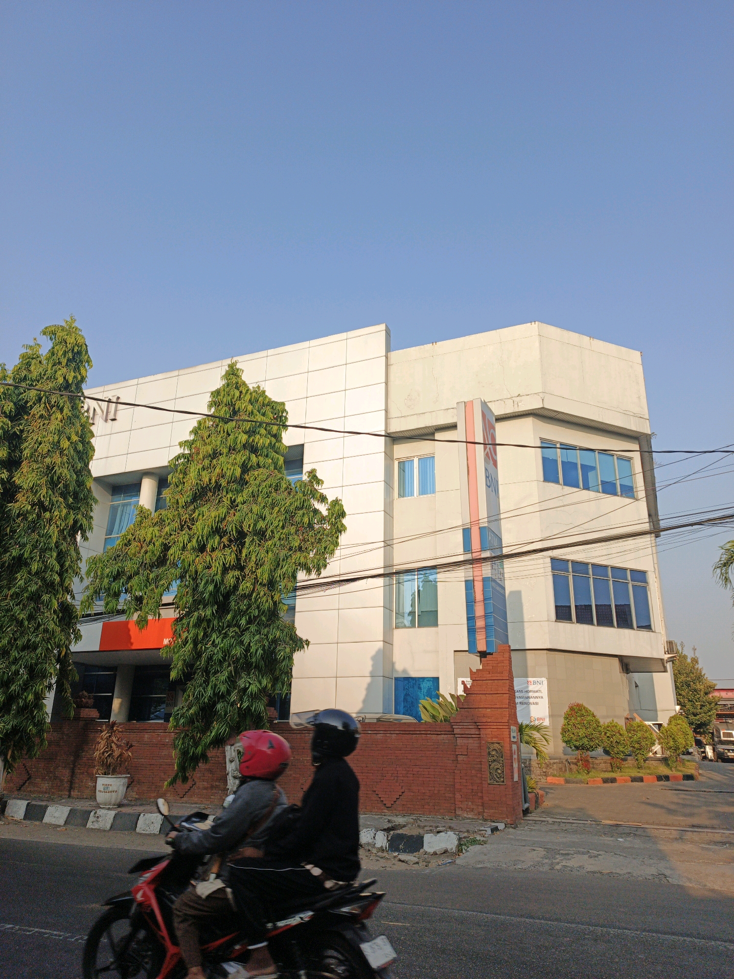 In Miji, Indonesia, a three-story building with a light-colored facade and blue-tinted windows stands under a clear blue sky. The building has a modern, angular design. A red and blue vertical sign with the "BNI" logo is attached to the front of the building. Two tall, dense green trees are positioned in front of the building on the left side. In the foreground, two people are riding a red motorcycle on a paved road. The rider in front wears a red helmet, and the passenger behind wears a black helmet. The motorcycle is in motion, indicated by a slight blur. A brick wall with decorative elements separates the building from the road. Power lines are visible crisscrossing in front of the building. The scene appears to be a street view in a commercial or business district.