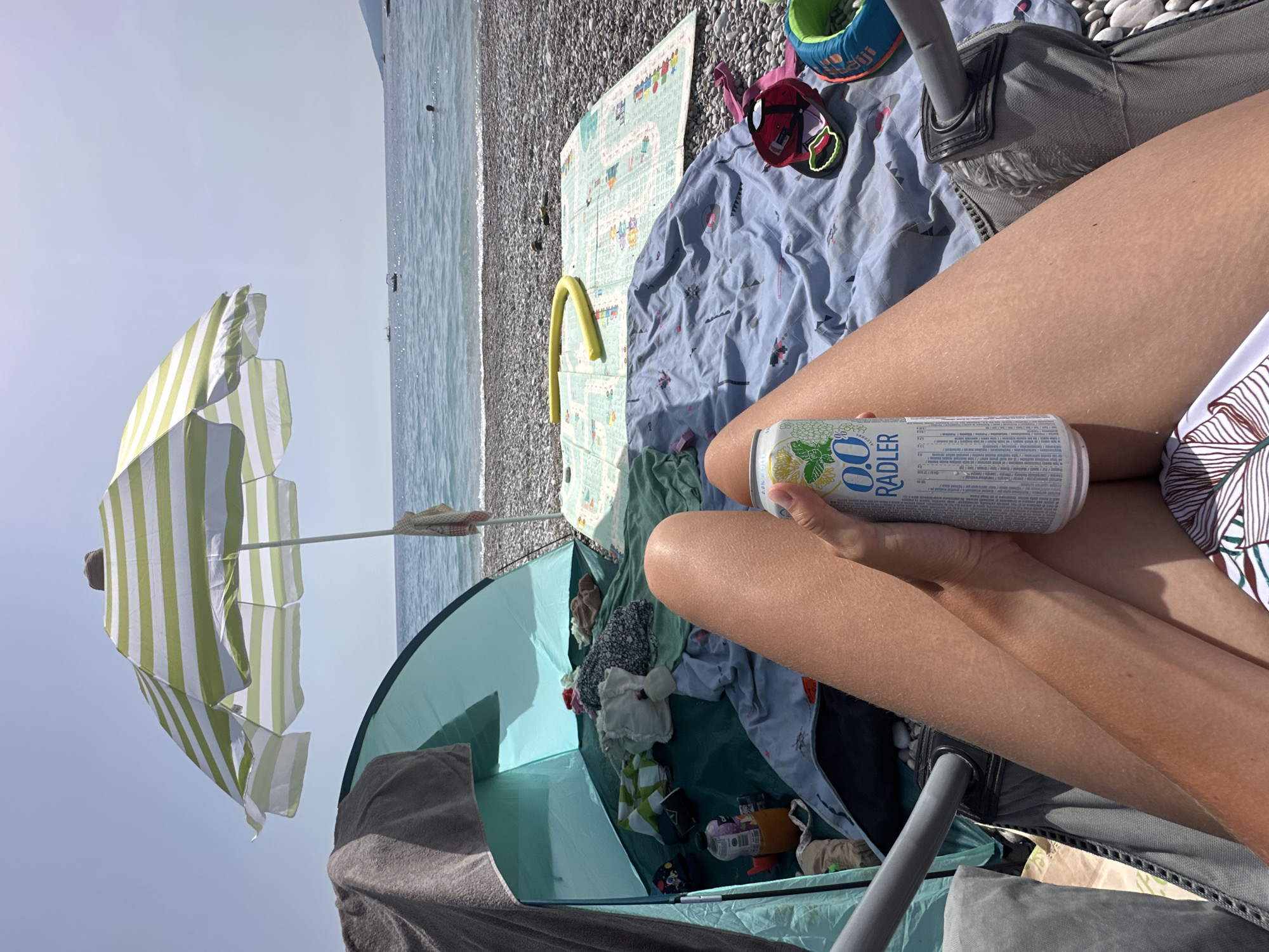 The image shows a beach scene, likely in Fethiye, Turkey, though without specific landmarks, confirmation is not possible. A person sits in a folding chair, holding a can of "0.0 Radler" drink. The scene includes: a beach umbrella with alternating white and light green stripes; a small blue beach tent containing clothing and toys; a beach blanket with a blue patterned cloth, a play mat, and children's items like a hat and shoes; a pebble beach, and the sea in the background. The composition is an elevated perspective, focusing on the person and their immediate surroundings.