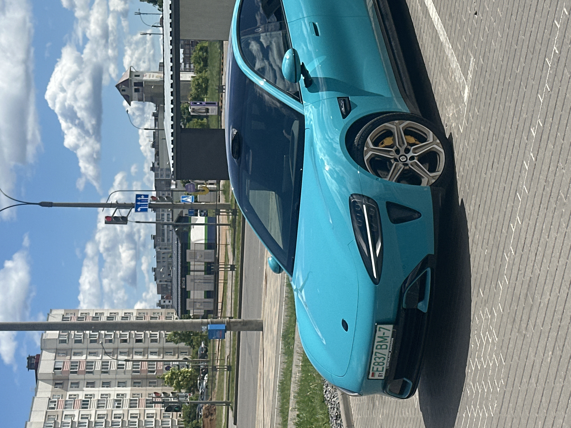 The image shows a turquoise-colored electric car parked in Бараўлянскі сельскі Савет, Belarus. The car has a license plate "FE837 BM-7". The foreground shows the vehicle and the parking spot and the background shows buildings, streetlights, traffic lights, and the sky. The photo was taken during the day under a bright sky with clouds.