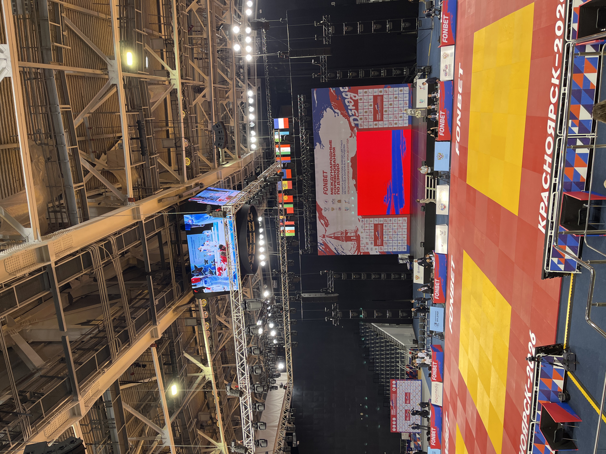 The image depicts an indoor sports arena, likely a judo or wrestling venue, in Krasnoyarsk, Russia, as indicated by the text "КРАСНОЯРСК-2026" visible on the mat. The arena features a large red and yellow competition mat, with a yellow central area. In the background, a large LED screen displays a graphic of a lighthouse and text, and above it, a smaller screen shows footage of athletes. Rows of empty blue seats are visible in the stands. The ceiling is a complex structure of metal beams, lighting rigs, and speakers. Several flags of different nations are hung across the upper part of the stage area. In the foreground, there are judges' tables with officials seated, and various branding displays for "FONBET." The overall scene suggests a sporting event is either about to begin or has recently concluded, with the focus on the competition area and supporting infrastructure.