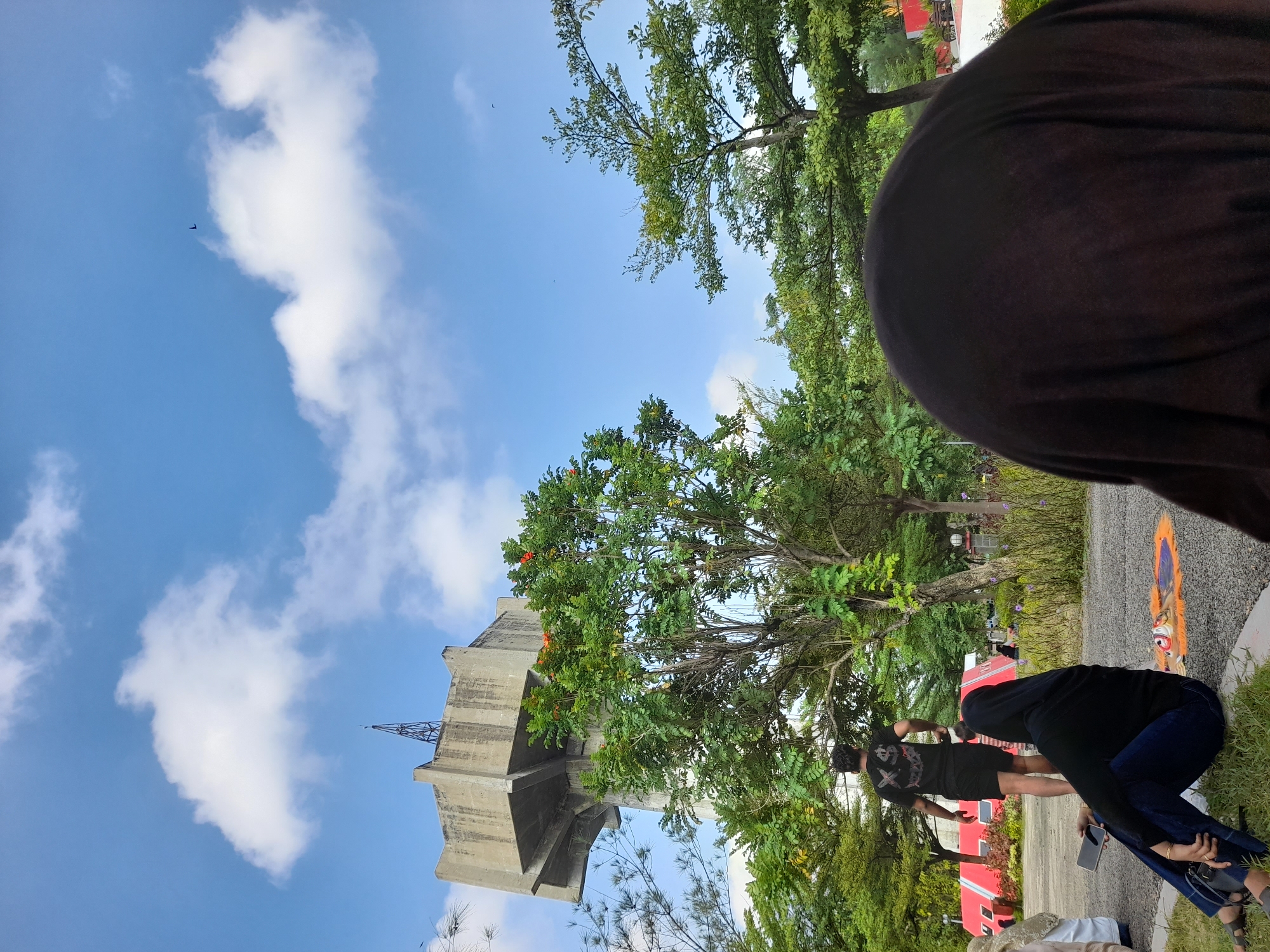 The image depicts an outdoor scene in Purwodadi, Indonesia, likely within a park or recreational area, given the presence of trees, green spaces, and a concrete structure that resembles a water tower. In the foreground, a person wearing a black hijab and blue jeans is seated on the ground, holding a smartphone. To their left, another person in a black t-shirt and shorts is walking away. In the background, the concrete water tower stands tall against a blue sky with scattered white clouds. Lush green trees and foliage surround the area. The overall impression is that of a leisurely day in a public space.