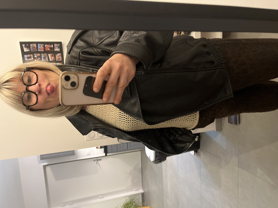 A woman takes a selfie in a mirror, sticking out her tongue. She wears black-framed glasses and has blonde hair. She's wearing a cream-colored, knitted top and a black leather jacket. Leopard print pants are visible. Behind her is a gray door and light gray tiles. A photo collage hangs on the wall above her head. The room is brightly lit.