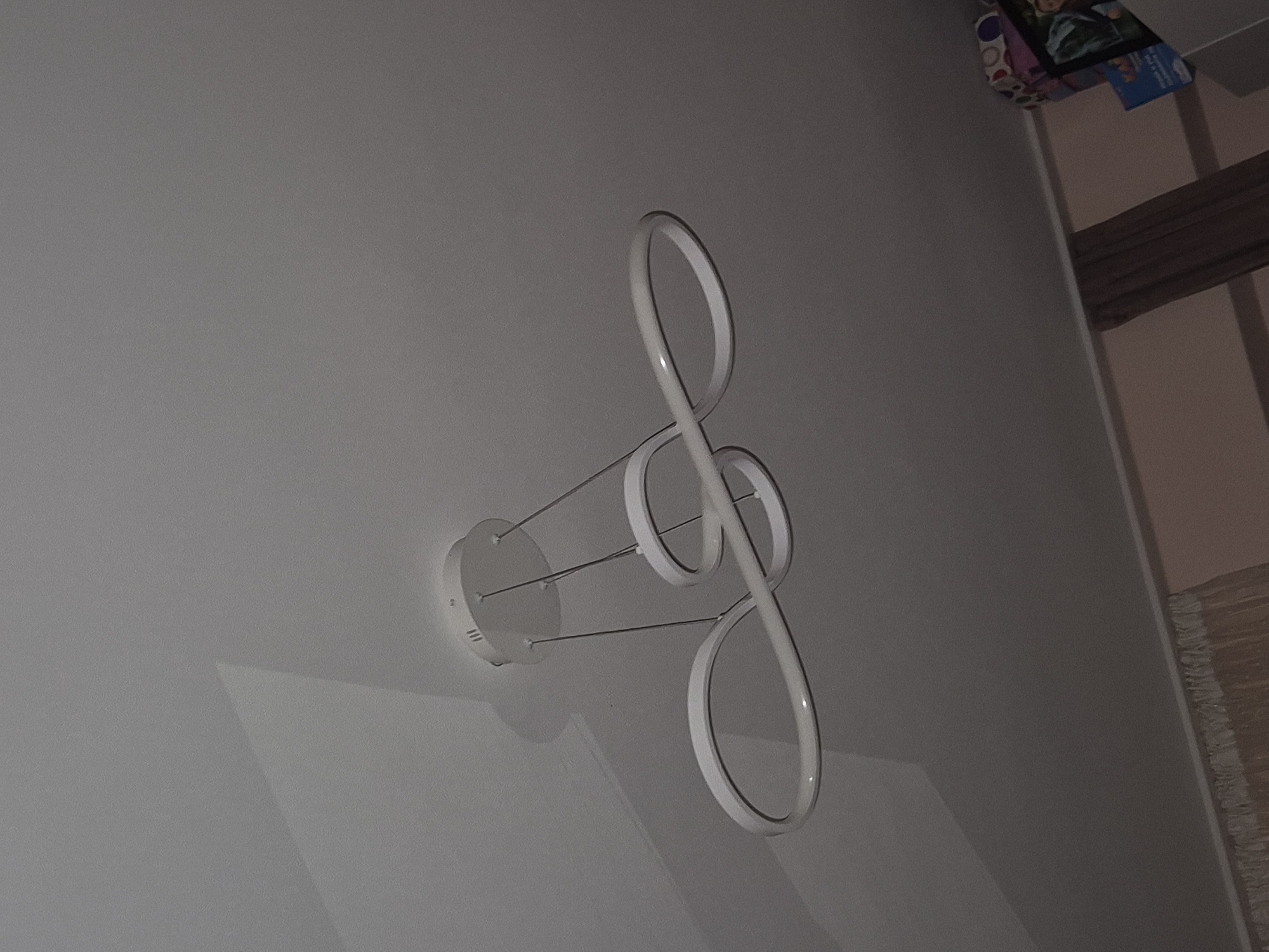 An indoor shot depicts a modern, white chandelier suspended from a textured ceiling. The chandelier features three interwoven, infinity-shaped loops, with thin metal wires connecting them to a circular mounting plate on the ceiling. Shadows cast by the chandelier are visible on the ceiling surface. The upper part of the image shows the ceiling, which appears to be a neutral color and slightly textured. The lower portion of the image includes a glimpse of a wall with curtains and some stacked boxes or decorative items, suggesting a domestic setting.