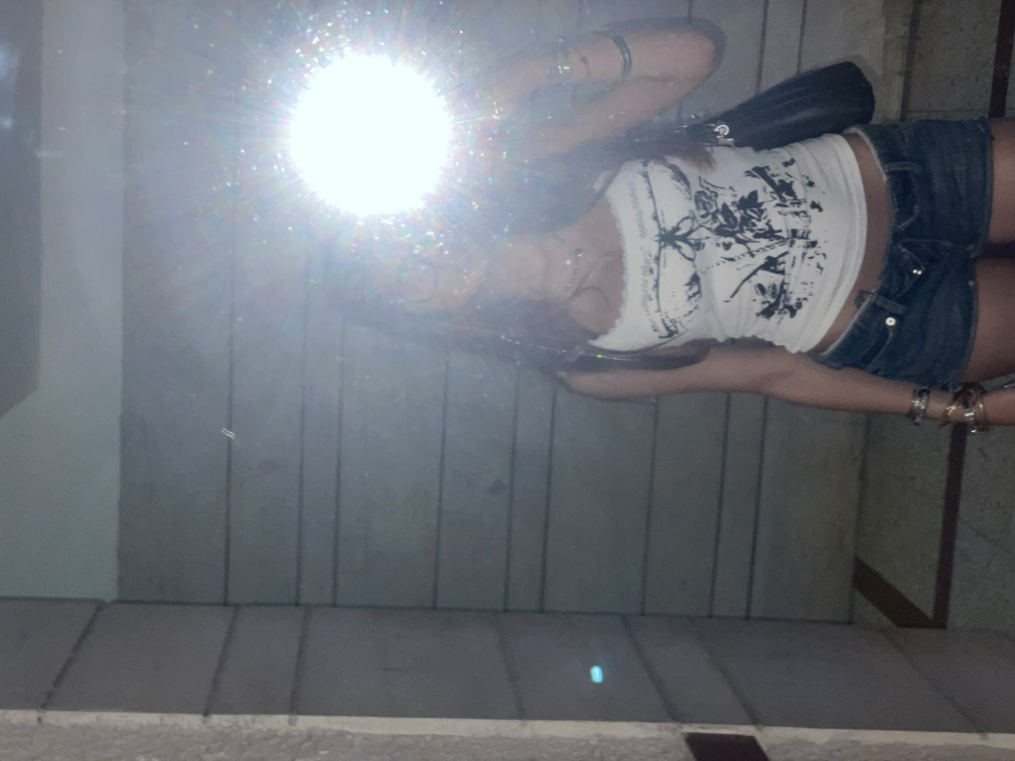 A young woman with long, auburn hair and glasses poses in a reflection in what appears to be a tiled hallway or doorway. She is wearing a white tank top with a black graphic print and denim shorts. She has several bracelets on her left wrist and is carrying a dark-colored purse. A bright light source, possibly a flash, is directly in front of her face, creating a strong lens flare and obscuring her features. The walls in the background are tiled with horizontal lines. The image is underexposed, with the flash significantly overexposing the central area. The environment suggests an indoor location, possibly in Petah Tikva, Israel, given the context provided.