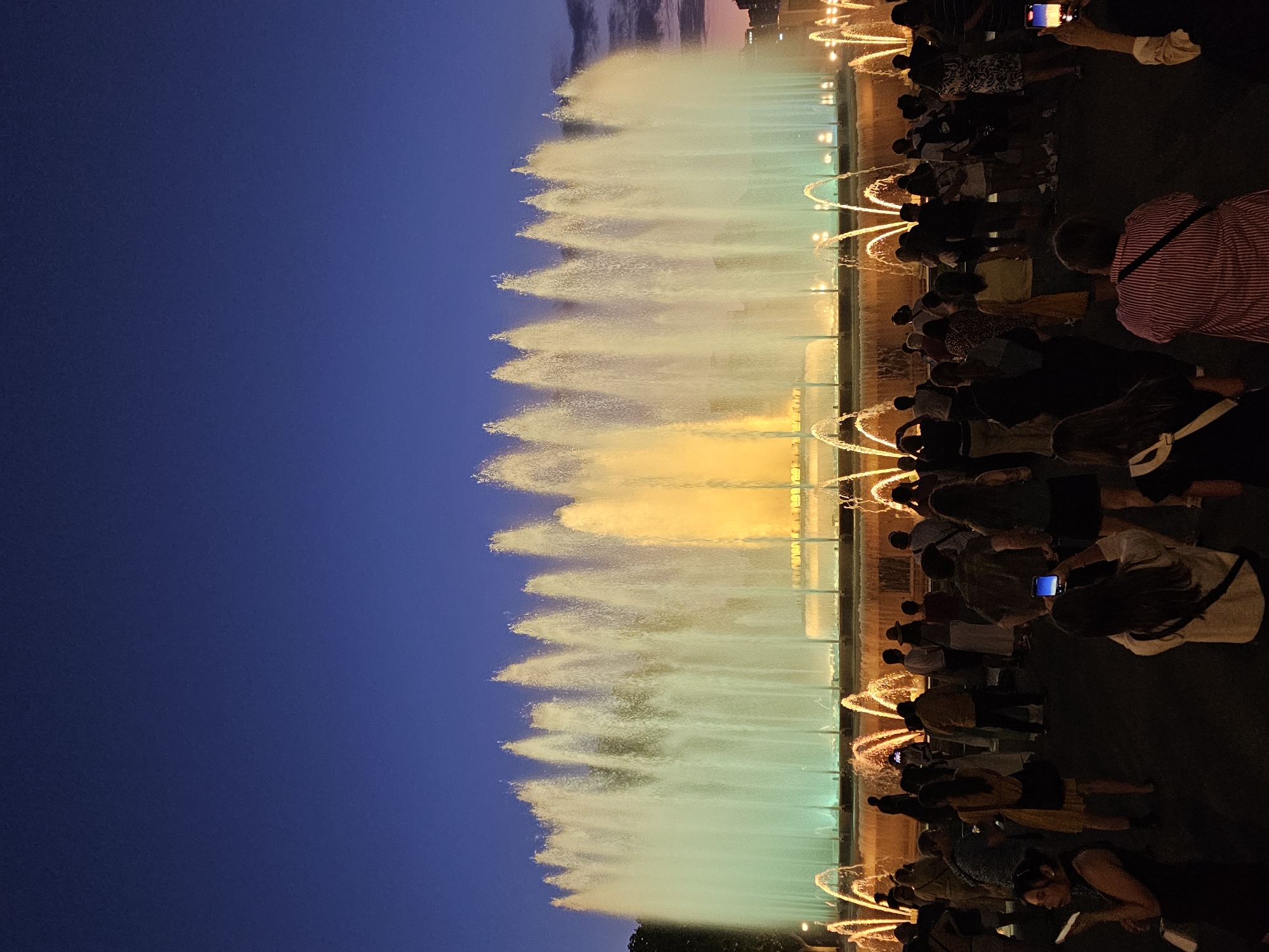 The image captures a nighttime view of the Magic Fountain of Montjuïc in Barcelona, Spain. A crowd of people is gathered in front of the fountain, illuminated by its colorful lights. The fountain itself is a central feature, with multiple jets of water creating a dynamic display. The water is lit with a combination of green and yellow hues, with some smaller arcs of orange light in the foreground. The sky above is a deep blue, indicating twilight or nighttime. The crowd appears to be diverse, with individuals standing and watching the fountain. Some people are holding up phones, likely to capture photos or videos of the spectacle. The environment suggests an outdoor public space, with a paved area for the audience.