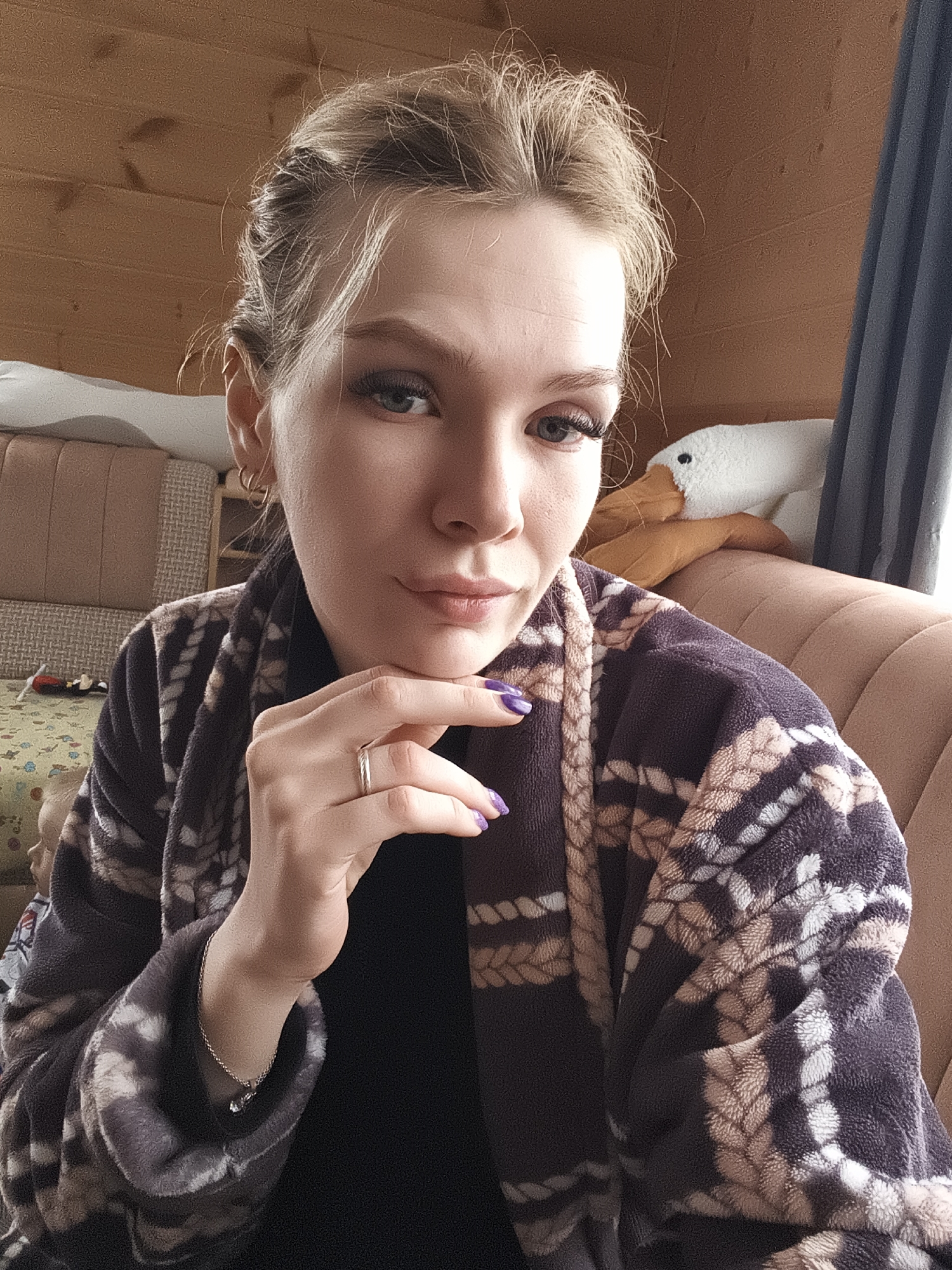 A young woman is sitting on a salmon-colored couch in a room with wood paneling. She is wearing a black top and a brown and beige robe with a rope-like pattern. Her hair is blonde and styled in an updo. She is wearing a ring on her left ring finger and a bracelet on her left wrist. Her nails are painted purple. She has light skin, blue eyes, and long eyelashes. She is resting her chin on her hand, and she is looking directly at the camera. Behind her, there is a plush white goose. The location is likely inside a home or residence, and it is possibly located in Русский Вожой, Russia.