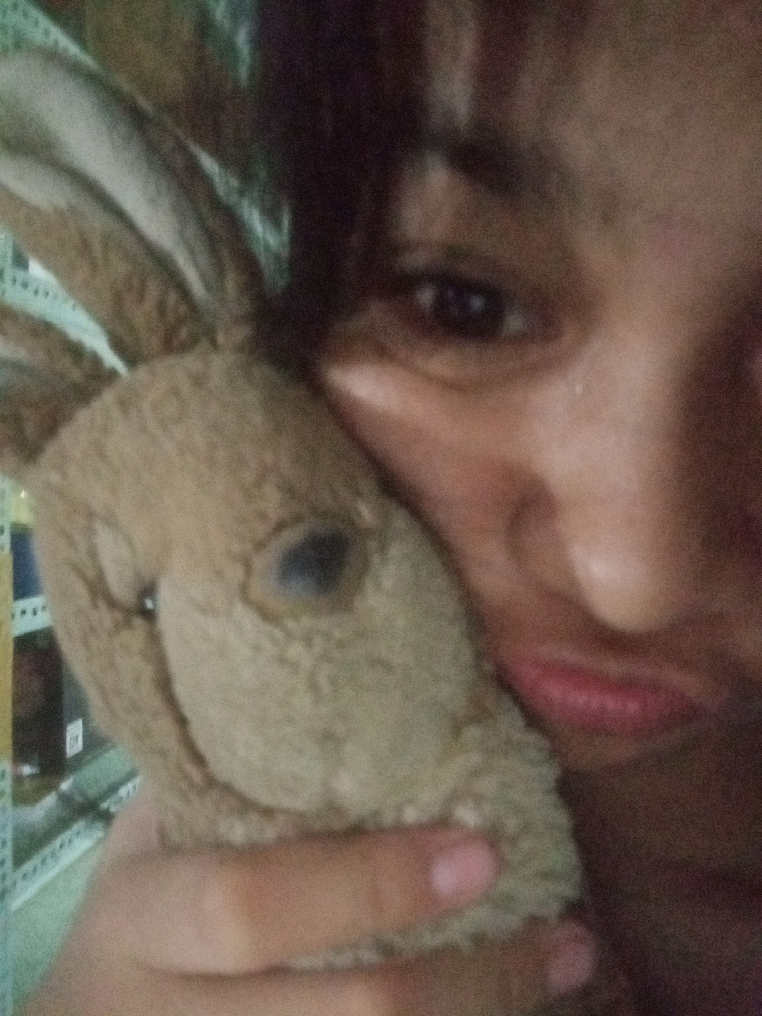A close-up, low-resolution, slightly blurry image shows a person holding a stuffed rabbit. The person's face is partially visible, with their right eye and mouth in focus. The stuffed rabbit's head is also in focus, appearing to be brown and fuzzy. The background is dark and out of focus, with some blurry shelves visible. The image is taken indoors, and the lighting is dim. There is no identifiable information about the location in the city of Badas, Indonesia.