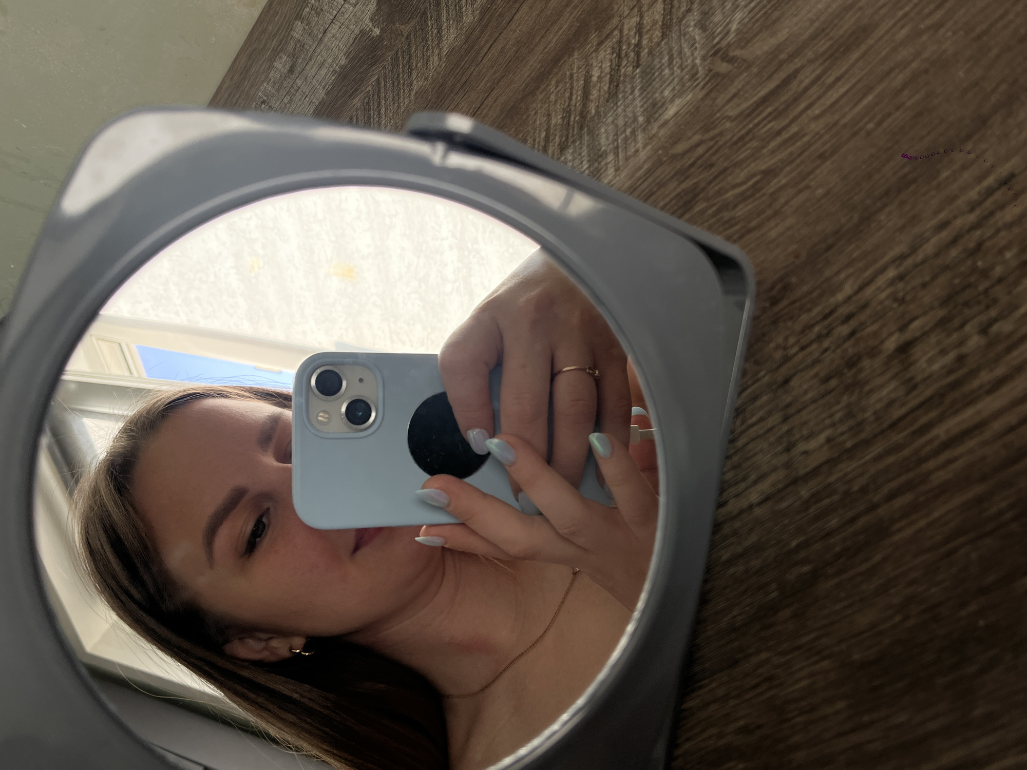 A young woman is reflected in a light-blue-framed mirror, holding a light-blue smartphone. The reflection shows part of her face, a gold earring, and a gold chain. The phone partially obscures her face, revealing the rear camera and a black sticker. Her hand with light-blue nails is also reflected. The mirror is placed on a surface that appears to be wood-grain. The location is not discernible from the image. It cannot be determined that the image was taken in Izhevsk, Russia.