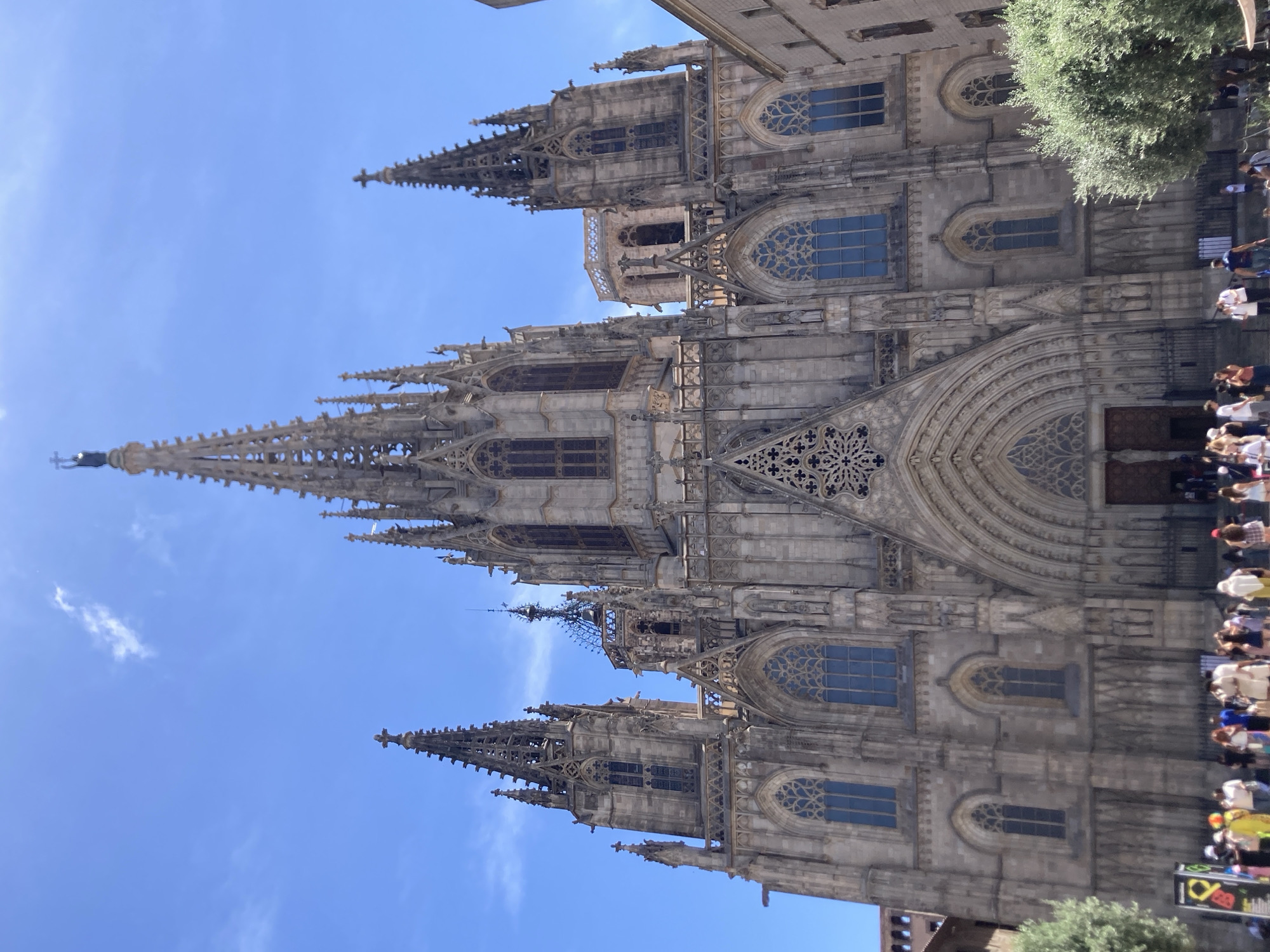 The image shows the Barcelona Cathedral in Barcelona, Spain, under a clear blue sky. The grand Gothic facade of the cathedral dominates the frame, with its intricate stonework, towering spires, and arched entrances. Several people are visible in the foreground, gathered in front of the cathedral, some appearing to be tourists. There are trees to the left and right of the cathedral, suggesting an outdoor public space. The overall scene depicts a sunny day at a prominent landmark in Barcelona.