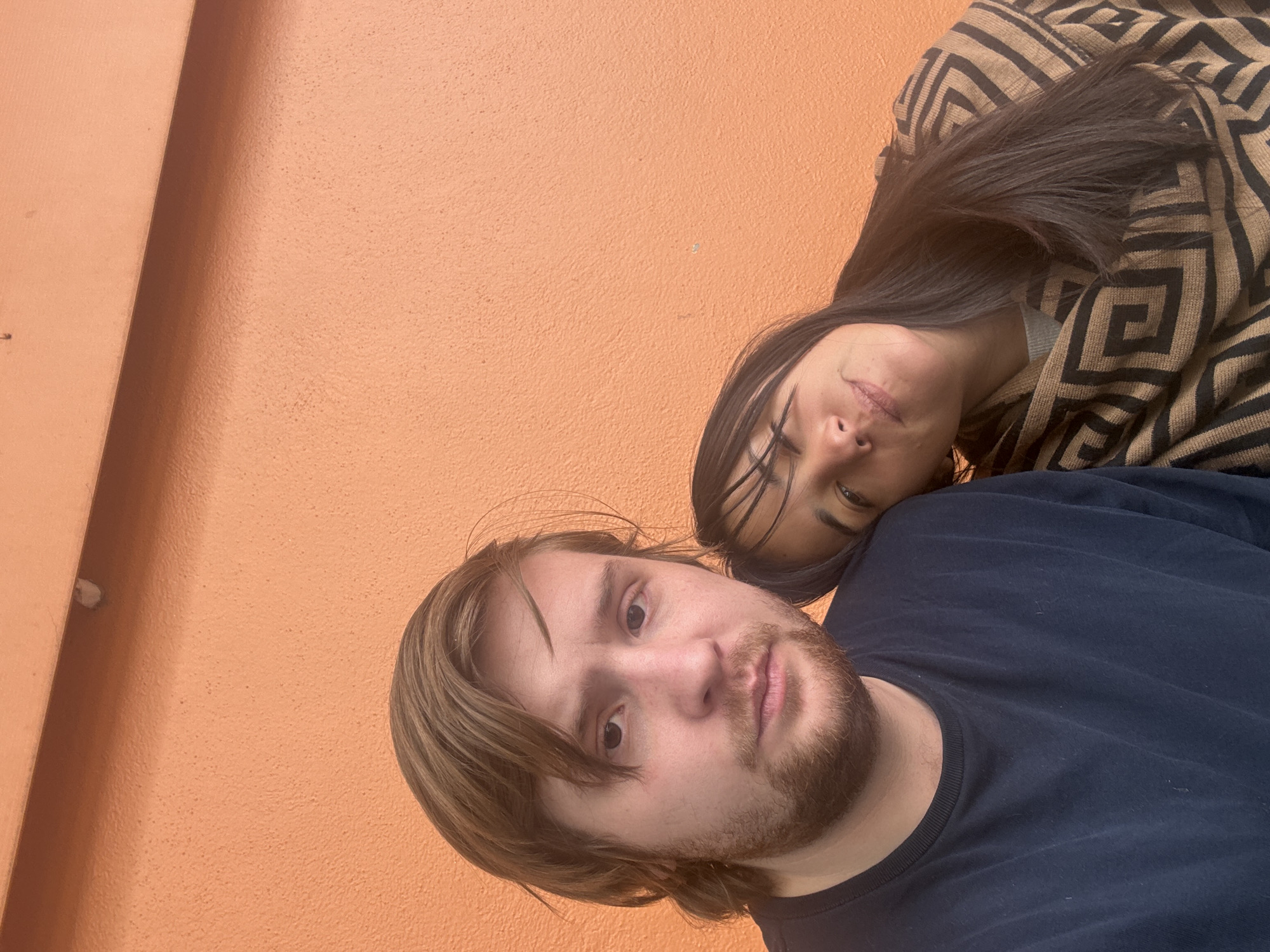 A selfie shows a man with light brown hair, a beard, and a dark blue t-shirt. A woman with long, dark hair and a patterned brown and black top is partially visible behind him. The background is a smooth, orange wall. The location is likely indoors. It is not possible to determine the exact location in Huixquilucan de Degollado, Mexico from the image.