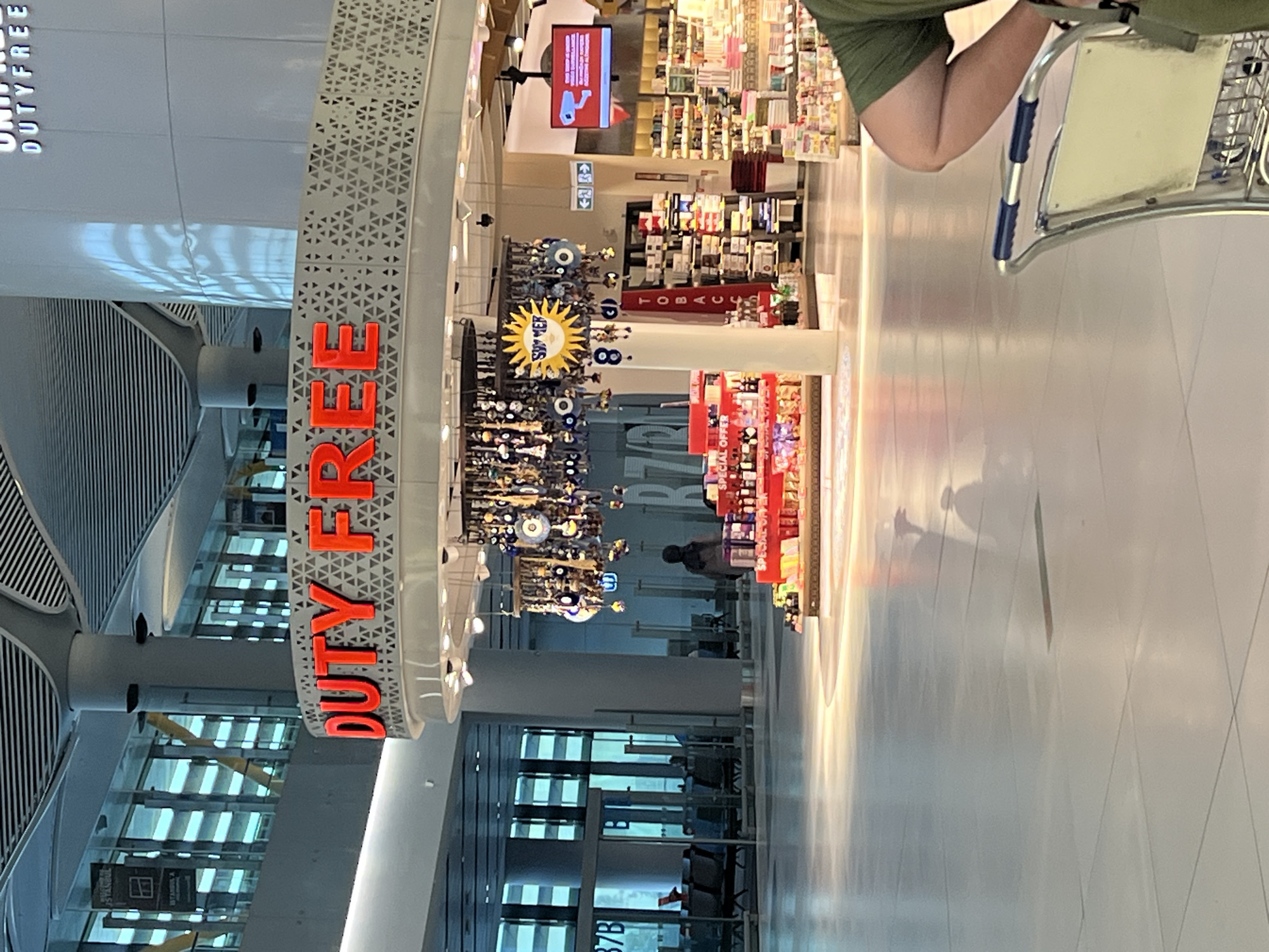 The image shows an interior view of a modern airport terminal, likely at Istanbul Airport in Arnavutköy, Turkey. The prominent "DUTY FREE" signage in bright red letters is displayed on a curved structure above a retail display. The display features various products, including bottles and boxes, with some labeled "SPECIAL OFFER." A decorative chandelier-like object hangs above the display, adorned with numerous small charms, including evil eye amulets. A white pillar stands near the display, featuring a sun graphic with the word "SUMMER" and the number "8" below it, along with vertical text that appears to read "TOBACCO." In the background, rows of merchandise shelves are visible, typical of a duty-free shop. Through large glass windows in the background, seating areas with people waiting can be seen, along with departure gate numbers, such as "37B." The floor is polished and reflects the ambient lighting. A person's arm and shoulder, wearing a green t-shirt, are visible in the lower right corner, partially obscuring a luggage trolley.