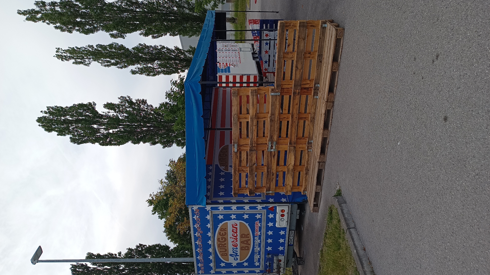 A food truck, branded as "BURGER American BAR," is parked outdoors under a blue canopy. The truck is decorated with an American flag motif and features stars and stripes. In front of the truck, a stack of wooden pallets is arranged, possibly for customer queuing or as a display. To the left of the truck, a shopping cart is visible. The scene is set in what appears to be a parking lot or an open area, with a gray asphalt surface, a strip of grass, and a concrete curb. Tall, slender trees are in the background, and the sky is overcast. A streetlamp is also present on the left side of the image. The food truck bears the license plate "M HO 2268". The location is identified as Unterhaching, Germany.