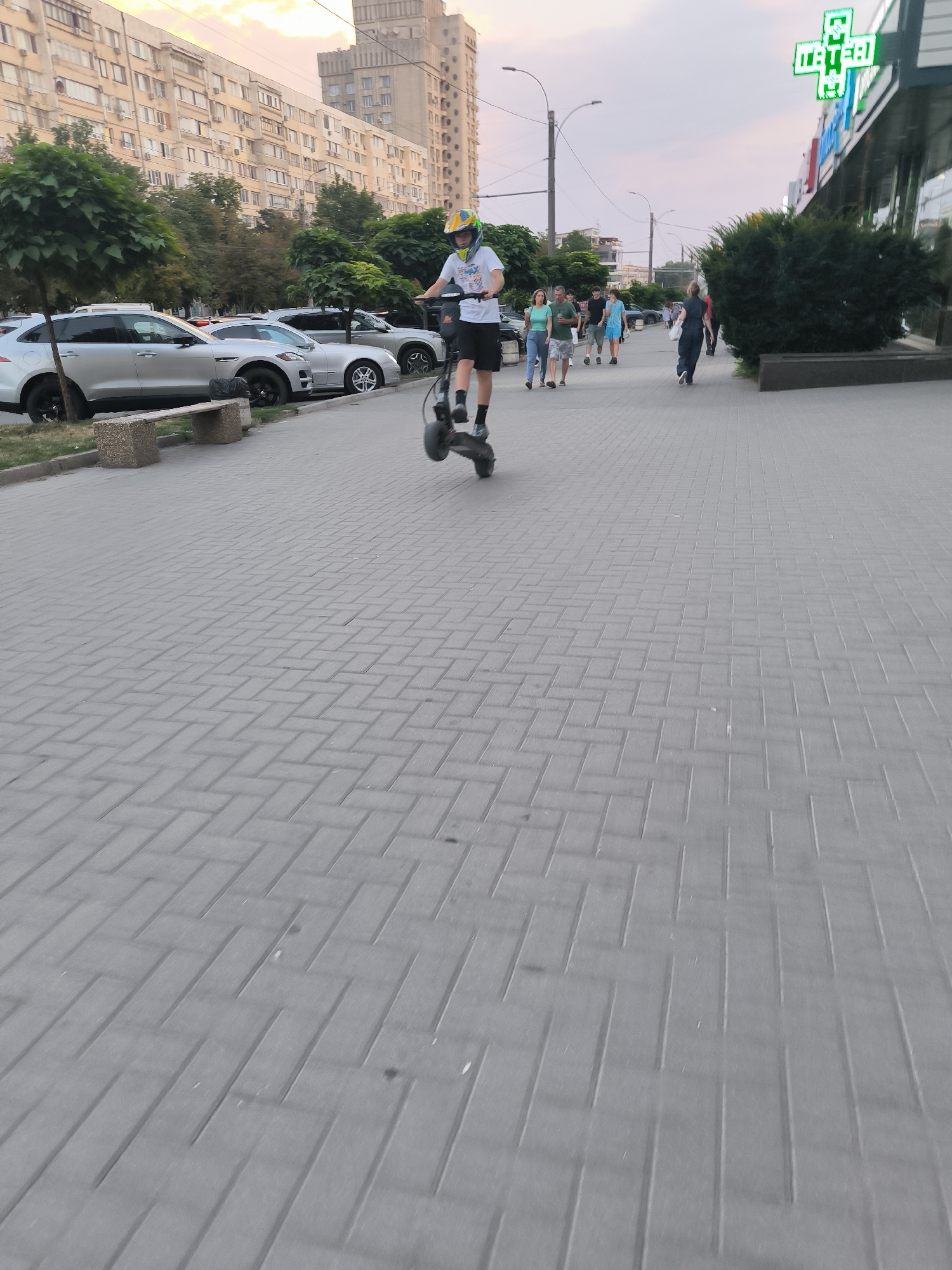 A young male on a black electric scooter with raised front wheel is in motion on a light gray patterned sidewalk. He is wearing a white t-shirt, black shorts, and a yellow helmet. In the background, several cars are parked along the street, and a line of people are walking on the sidewalk. To the right, a green illuminated sign with text "MATERNA" is visible above a building. The sky is pale with hints of pink and orange. The scene appears to be in Chișinău, Moldova, near the Materna pharmacy on Alexandru cel Bun street.
