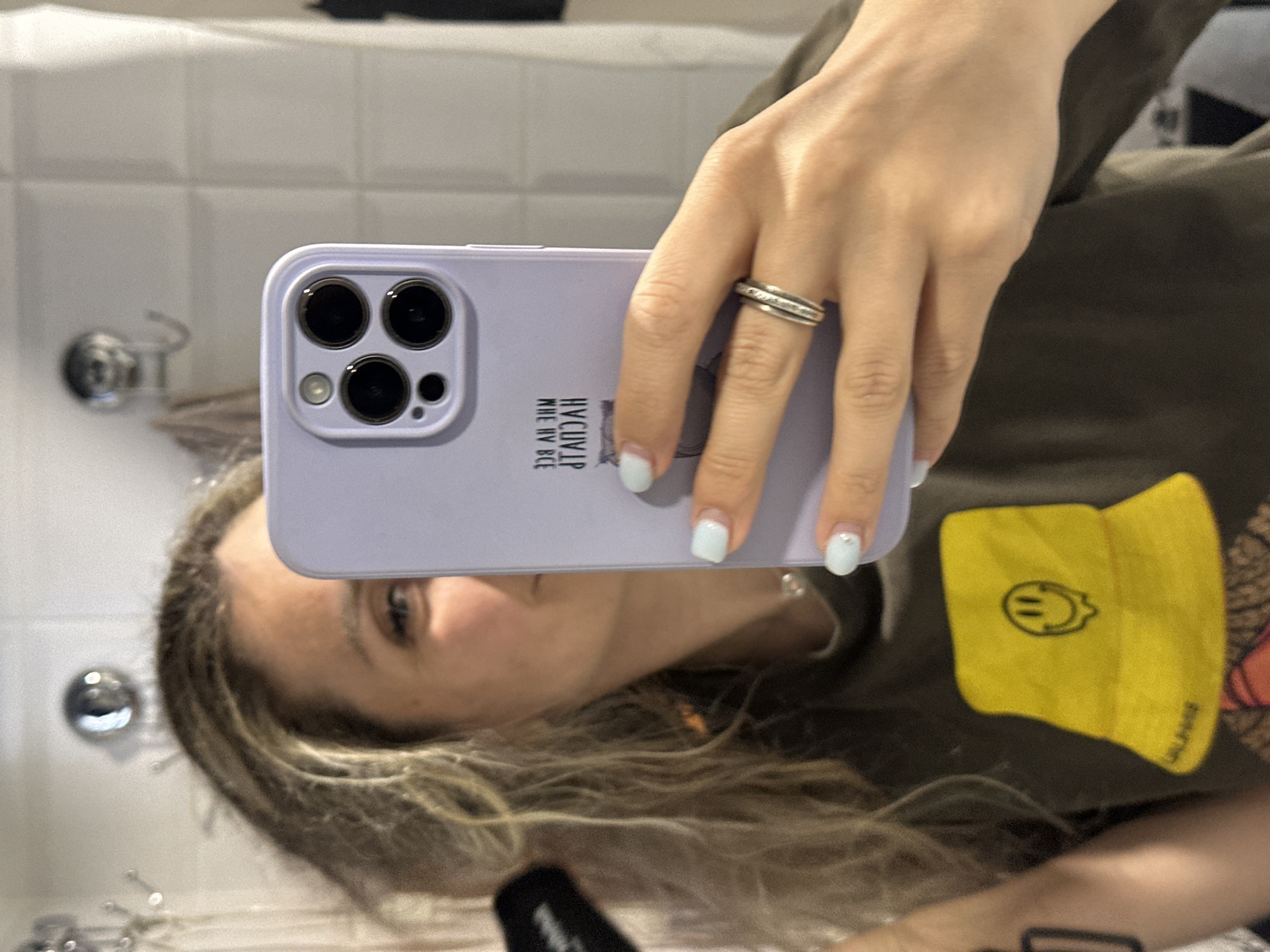 A person is taking a mirror selfie in a bathroom. The person has blonde hair and is wearing a dark green t-shirt with a yellow bucket hat graphic. They are holding a light purple phone with a triple camera lens. The bathroom has white tiled walls and a faucet is visible in the background. There is also a ring on the person's finger and light blue nail polish. The image appears to be taken indoors.