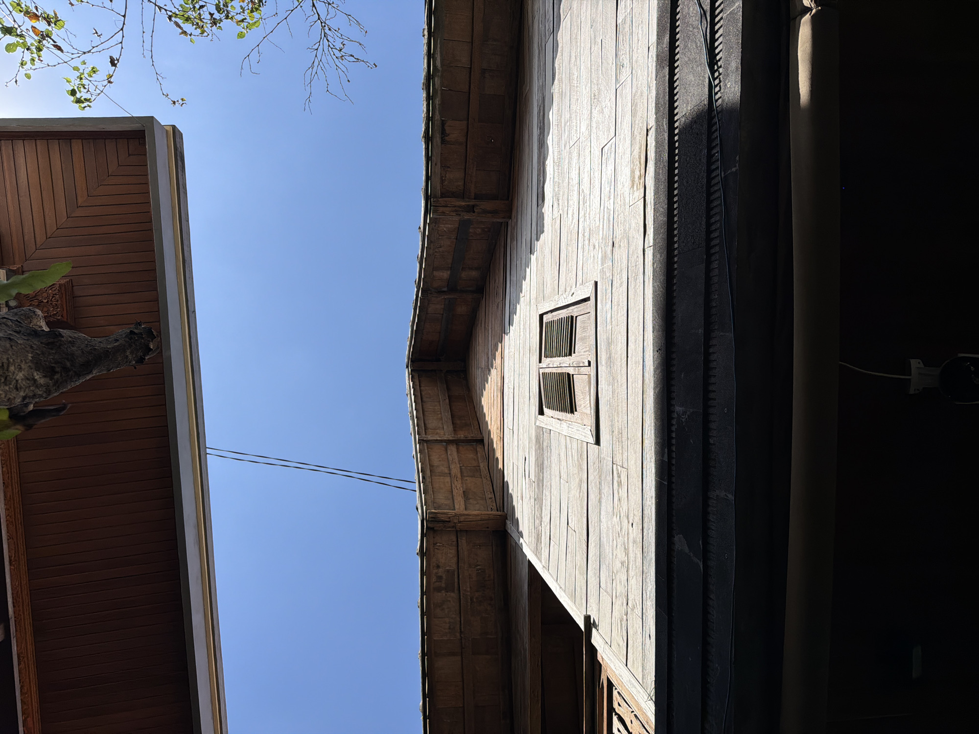 This upward-angled photograph, captured in North Kuta, Indonesia, depicts the upper facade of a weathered wooden building against a clear, bright blue sky. In the upper left, a textured, dark tree trunk with sparse green foliage partially frames a detailed brown wooden soffit, featuring a concentric square pattern. To the right, bare tree branches with a few green leaves extend into the sky. Two black utility wires traverse the sky diagonally from the upper left quadrant. The building's prominent feature is a gabled upper section constructed from horizontal wooden planks, which show signs of weathering and varying coloration. Centrally located on this section is a small, rectangular window fitted with closed, horizontally louvered wooden shutters. Below this, the building's exterior is clad in horizontally arranged, light-toned wooden boards. A dark, horizontal architectural band with a repeating pattern of small, regular indentations or carvings is visible beneath the wooden boards. The lower section of the image is predominantly in deep shadow, revealing a light-colored, cylindrical object spanning horizontally. In the bottom right corner of the shadowed area, a white electrical conduit and a small white fixture are discernible. No individuals are present in the scene.